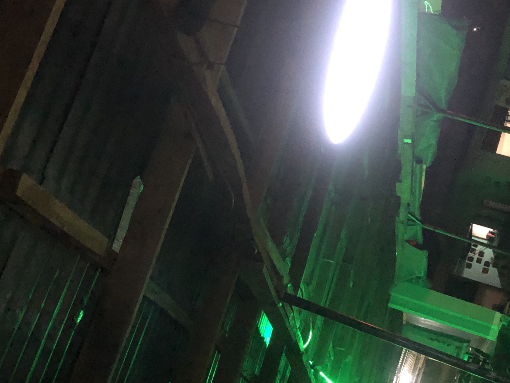 An exterior, eye-level, medium shot captures the underside of a rustic, open-air shelter with a corrugated metal roof supported by wooden beams. The shelter is illuminated by bright green LED strip lights, which cast a vibrant glow on the wooden structure and create a somewhat surreal atmosphere. To the right, a bright white circular light fixture emits a strong, diffused light that washes out a portion of the upper right corner of the image. The overall scene suggests a dimly lit outdoor space, possibly a market stall, a covered seating area, or an entertainment venue at night. The image is dark and grainy, with the green and white lights being the primary sources of illumination. The location is identified as Unknown, Unknown.