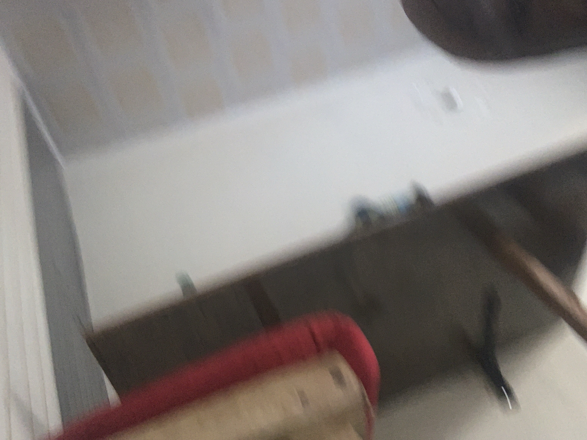 The image depicts an indoor scene, likely a room. In the foreground, a red upholstered object, possibly a cushion or seat, rests on a wooden surface. Behind this, a shelf is visible, sparsely decorated with a few indistinct items. The background consists of a wall and a ceiling. The lighting appears to be ambient, and the overall image is out of focus, suggesting a low-quality camera or a shaky hand. There is no indication of the specific location within Jauro Yinu, Nigeria, as the image lacks any recognizable landmarks or identifying features.