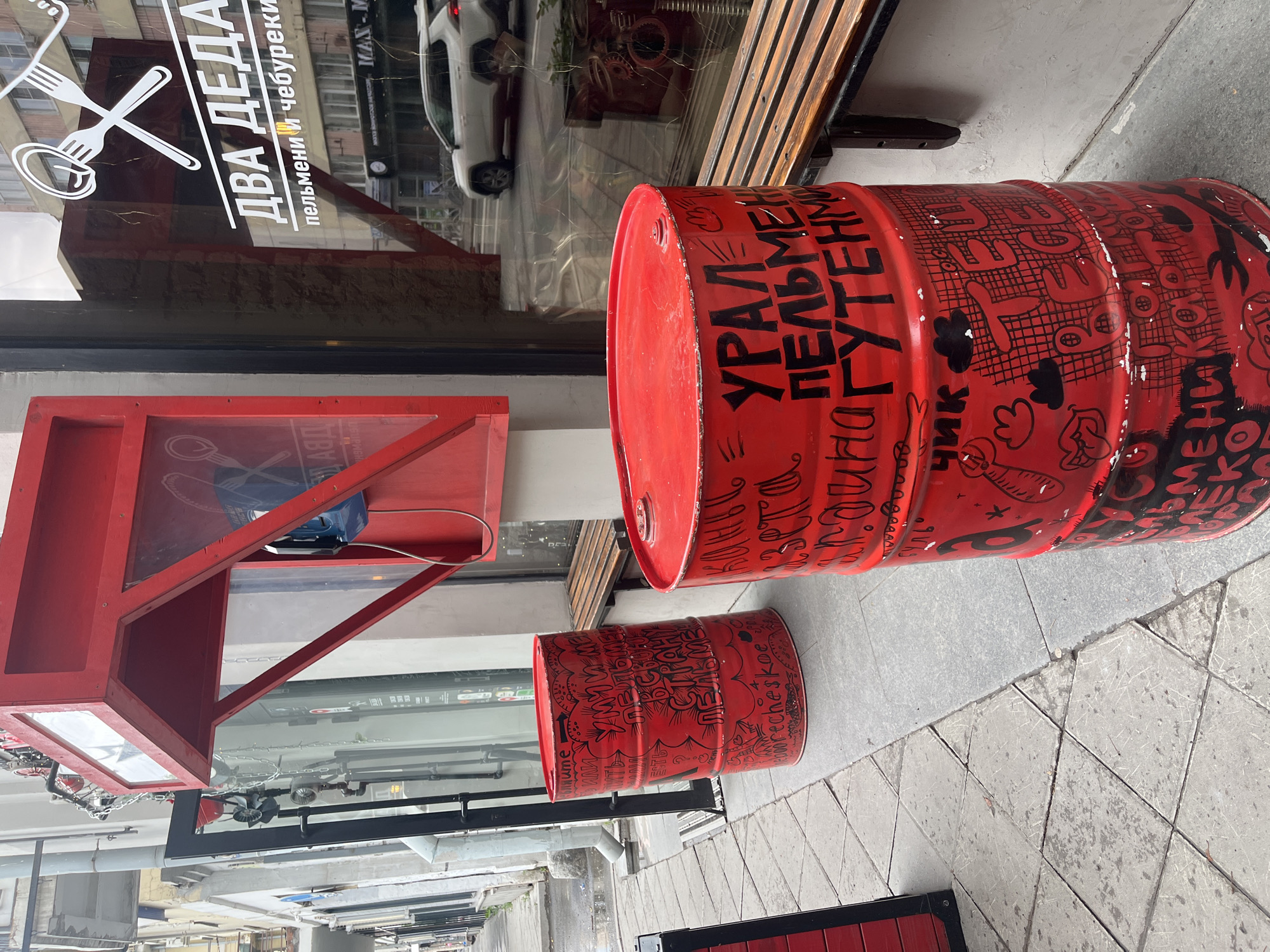 Here is a detailed description of the image:
The image captures a street-level scene in the city of Unknown, Unknown. Two red painted oil barrels are visible on the sidewalk, with bold black graffiti covering their surfaces. One barrel is noticeably larger than the other. Beside the barrels is a red, box-like structure, possibly an outdoor telephone booth or public service kiosk.
Reflections in the glass windows of a building behind the barrels reveal portions of the street, including parked cars and pedestrians. The storefront advertises a restaurant named "ДВА ДЕДА", which translates to "Two Grandfathers", specializing in dumplings and chebureki.
The surrounding environment suggests an urban setting with concrete sidewalks and typical city infrastructure. The overall scene suggests a mix of commercial activity and urban street art.