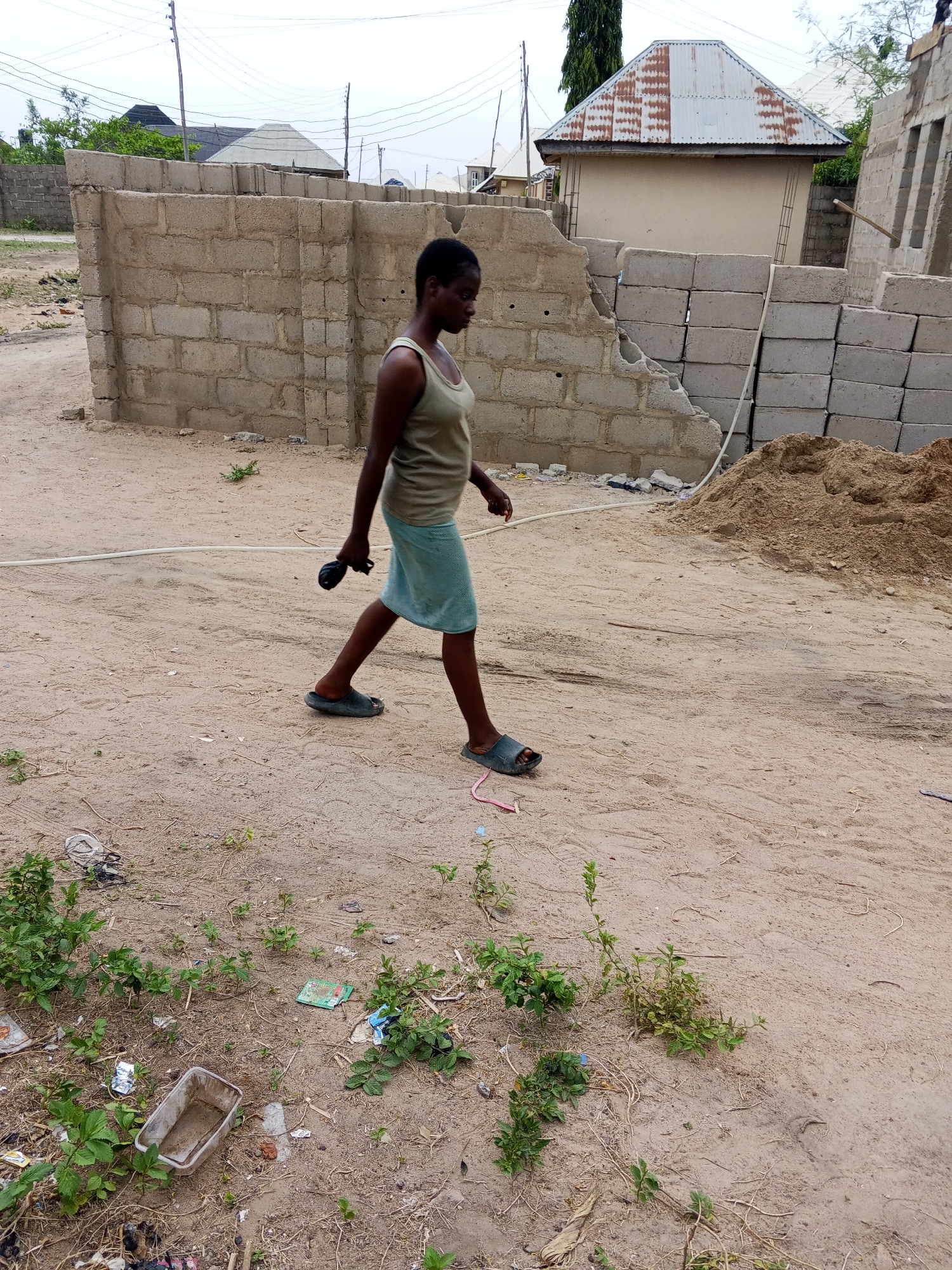 The image depicts a young woman walking from left to right across a sandy, unpaved area. She has dark skin and short, dark hair, and her gaze is directed forward. She is wearing a light-colored tank top, possibly olive green, and a light blue-green skirt or wrap. On her feet are dark slides or sandals. In her left hand, she holds a small, dark, cylindrical object.
The scene appears to be an outdoor, undeveloped or under-construction residential area. The ground is primarily dirt and sand, with scattered patches of small green weeds and various pieces of litter, including a clear plastic container, wrappers, and a pink ribbon-like object. A white hose or pipe runs across the mid-ground.
In the background, there are several concrete block walls in various states of construction or disrepair. To the left, a partially broken wall stands, with more completed block walls and buildings visible behind it. To the right, a large stack of concrete blocks is piled next to a mound of sand or dirt. Further back, a small, light-colored building with a distinct corrugated metal roof is prominent, flanked by another building with a dark roof. Overhead, power lines and utility poles are visible against a bright, overcast sky.
The activity is simply the woman walking. The lighting suggests it is daytime, possibly with an overcast sky. There is no visible text in the image.
