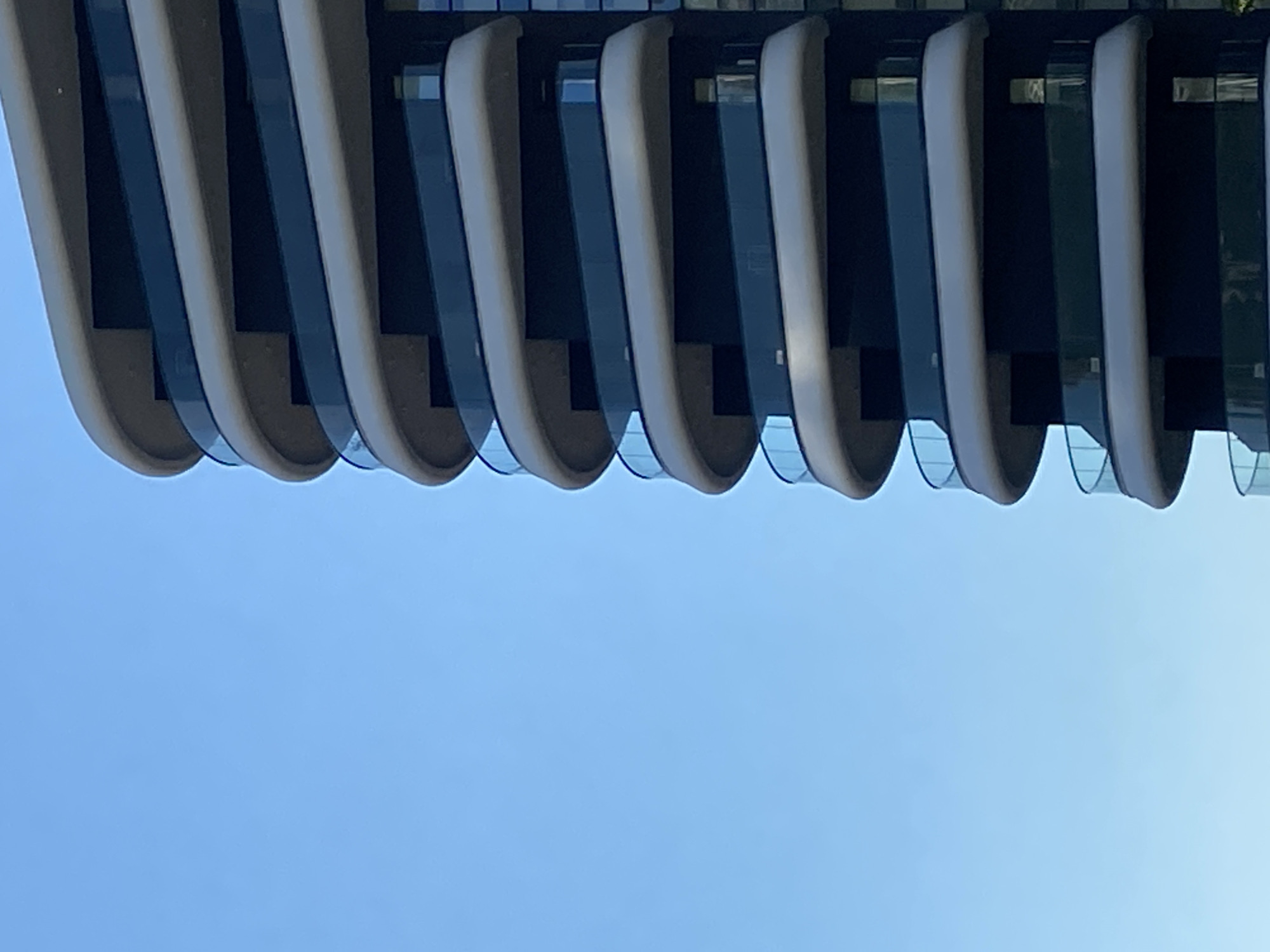 A low-angle, upward-facing, and slightly rotated perspective depicts a portion of a modern building in Boreti, Montenegro. The building's architectural facade is characterized by vertically oriented, curved structural elements in a light gray color. These elements are spaced evenly apart, revealing dark-colored glass or panels in the gaps between them. The sky, a uniform light blue color, serves as the background. No people or discernible activities are visible. The focus is on the building's design against the clear sky.