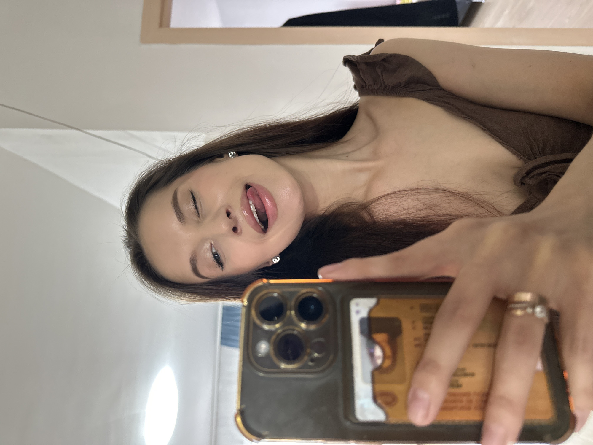 A young woman takes a selfie with her phone in a brightly lit room with white walls. She has long brown hair and is wearing a brown top. Her tongue is sticking out and one eye is closed. The phone is a newer model with multiple lenses and a gold-colored case, possibly an iPhone. The location is not identifiable as Yekaterinburg, Russia based on the image content.
