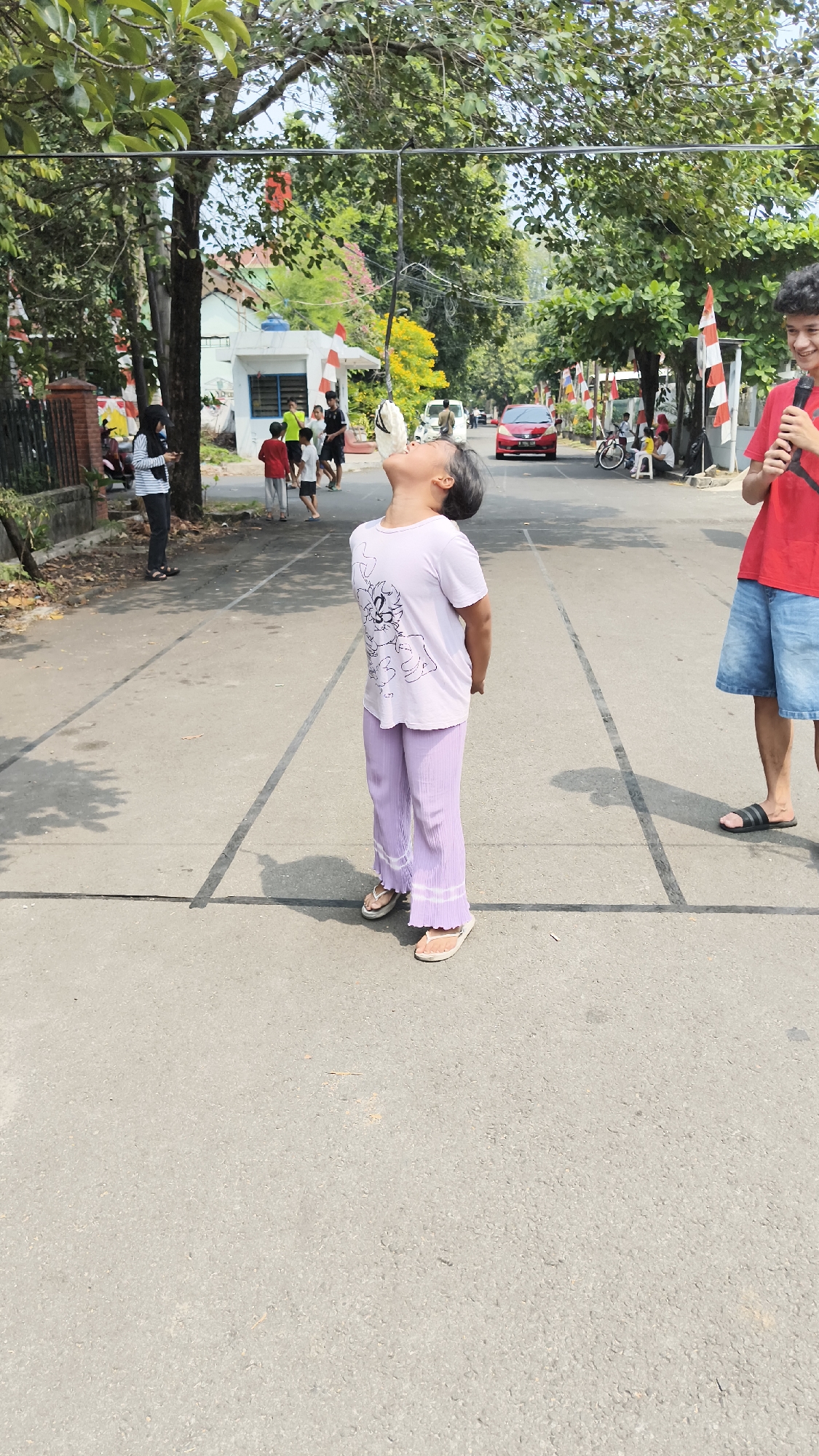 In South Jakarta, Indonesia, a young girl is participating in a traditional Indonesian Independence Day game, likely a cracker-eating contest. She is standing in the middle of a street, looking up at a cracker suspended from a string. She is wearing a white t-shirt with a graphic design and light purple pants. Her hands are behind her back. A man in a red shirt and blue shorts stands to her right, holding a microphone and smiling. In the background, there are trees, houses, parked cars, and other people, some of whom are watching the game. Indonesian flags are visible on flagpoles. The road is marked with white lines. The scene is set on a sunny day.
