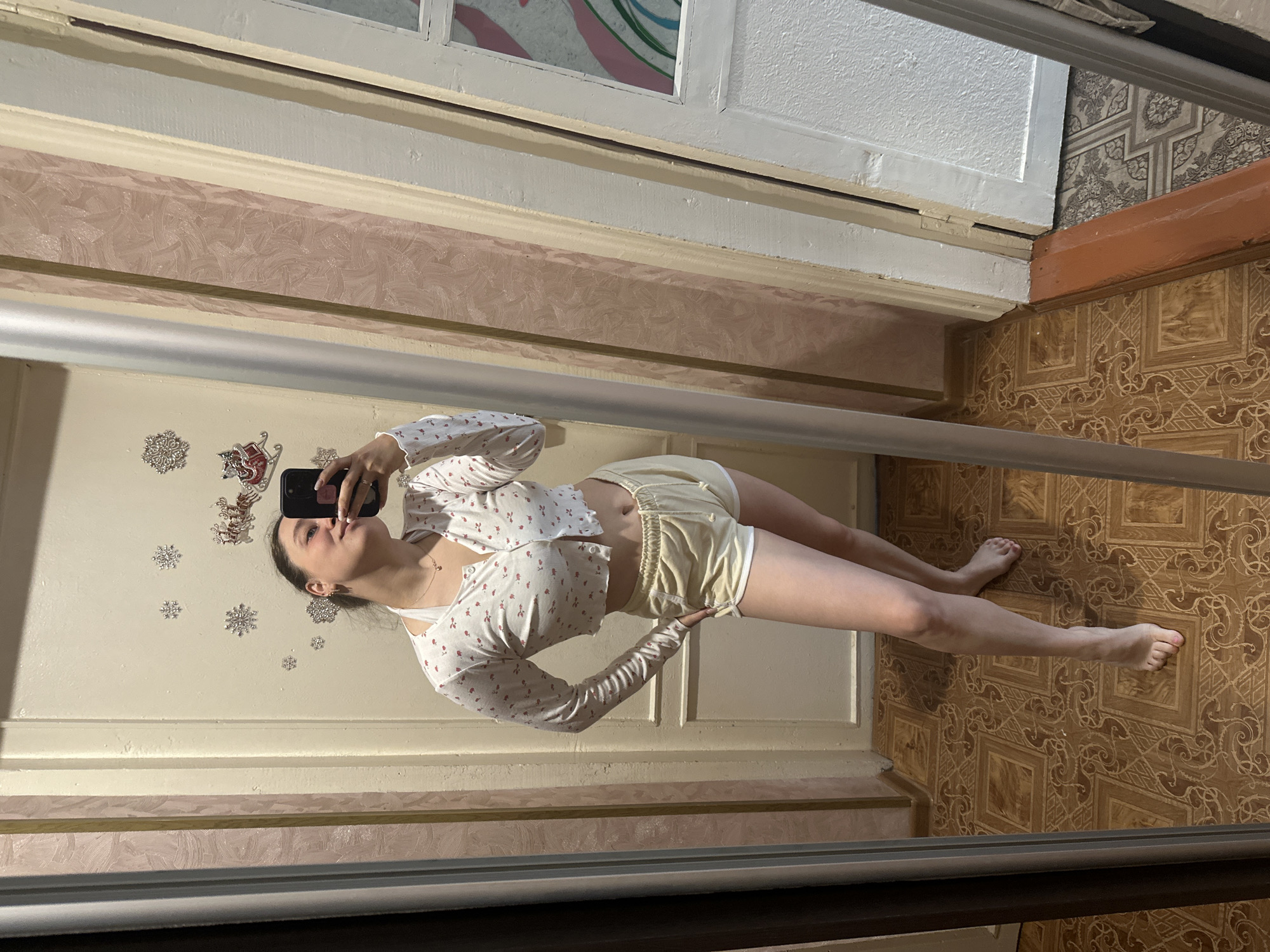 In a full-length mirror reflection, a young woman stands in what appears to be a residential interior. She is wearing a white cropped long-sleeved cardigan with a floral pattern and a white bralette underneath, paired with light yellow shorts. Her right hand is holding a smartphone, angled to capture the selfie, and she is leaning slightly forward, puckering her lips. The background shows a wall with light-colored paneling and wallpaper, adorned with snowflake and reindeer decals, suggesting a festive or seasonal decoration. The floor is tiled with a brown, intricate pattern. The scene is lit by an unseen light source, creating reflections in the mirror and casting soft shadows. The location is specified as Yaroslavka, Russia.