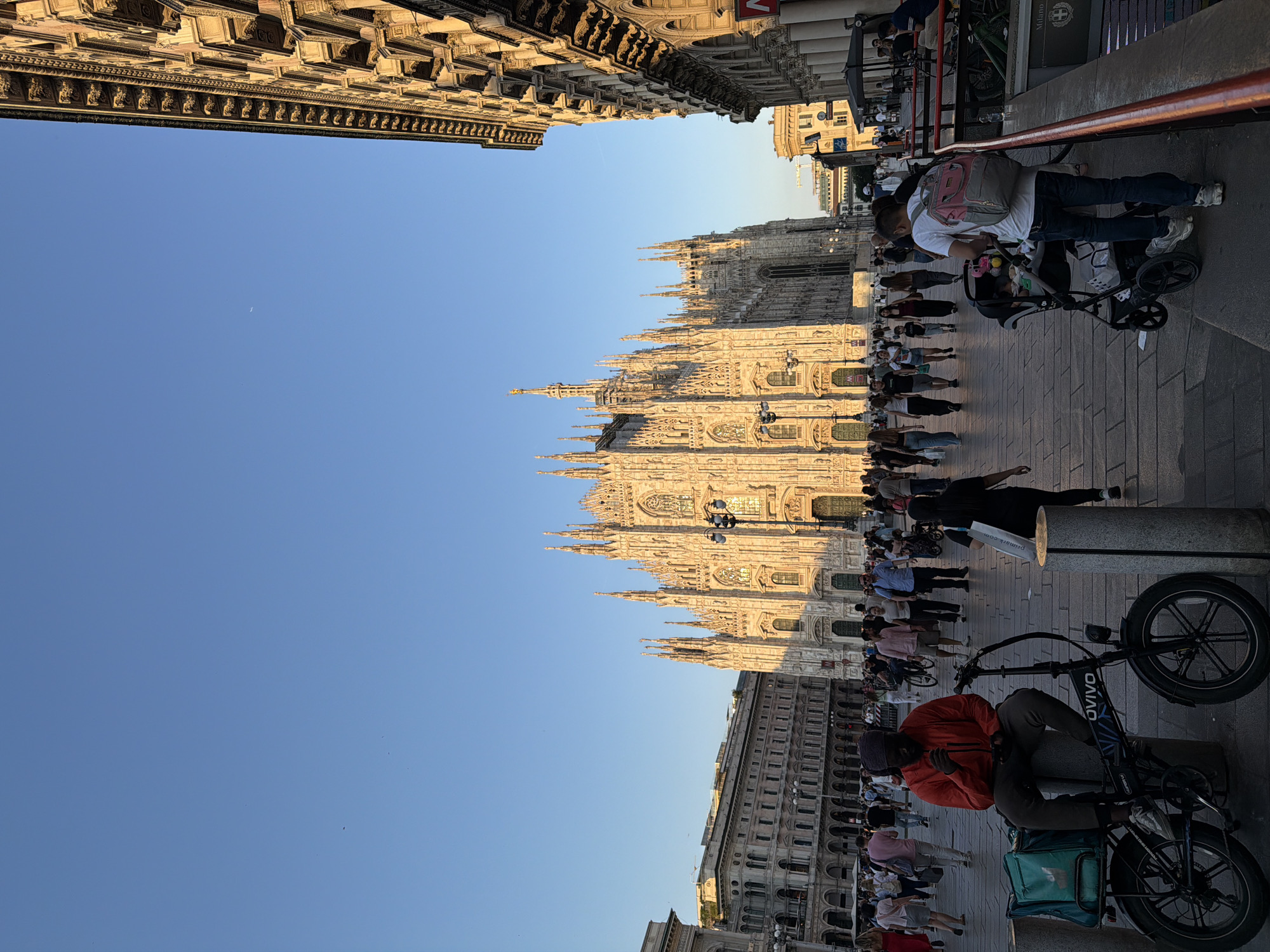 The image depicts Milan Cathedral, Italy, with a throng of people walking around. The shot is taken from street level. There are buildings around the cathedral. The sky is clear. People are walking, and some are pushing strollers. There are electric bicycles parked nearby.