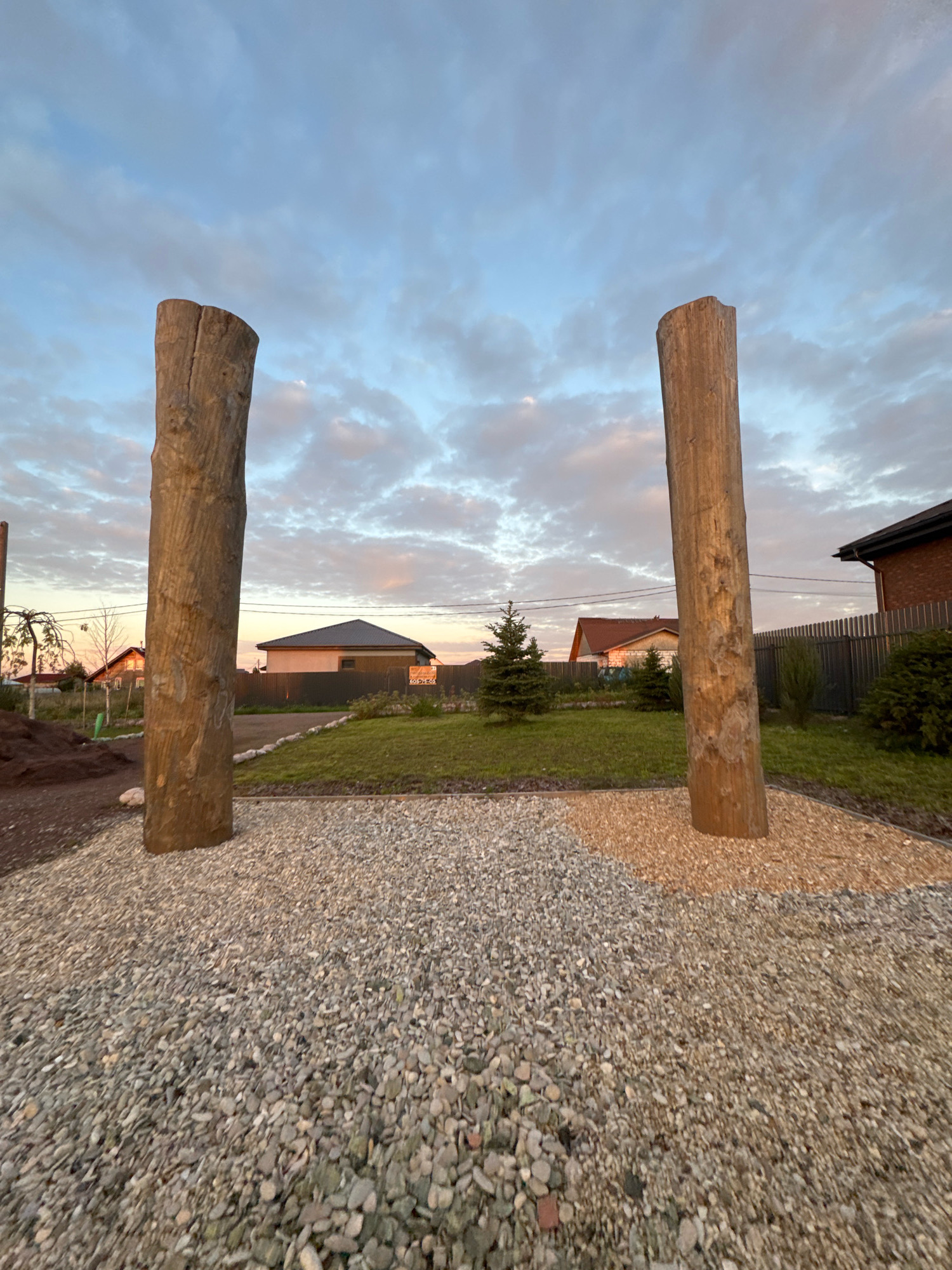 The image depicts an outdoor scene in Timmolovo, Russia, during what appears to be sunset. Two large, roughly hewn wooden posts stand vertically in the foreground, separated by a path covered in grey and orange gravel. Beyond the posts, a manicured lawn stretches out, punctuated by a small evergreen tree. In the background, residential buildings with dark roofs are visible, along with a metal fence and some vegetation. The sky above is filled with clouds, tinged with the colors of the setting sun. There are no people visible in the image.