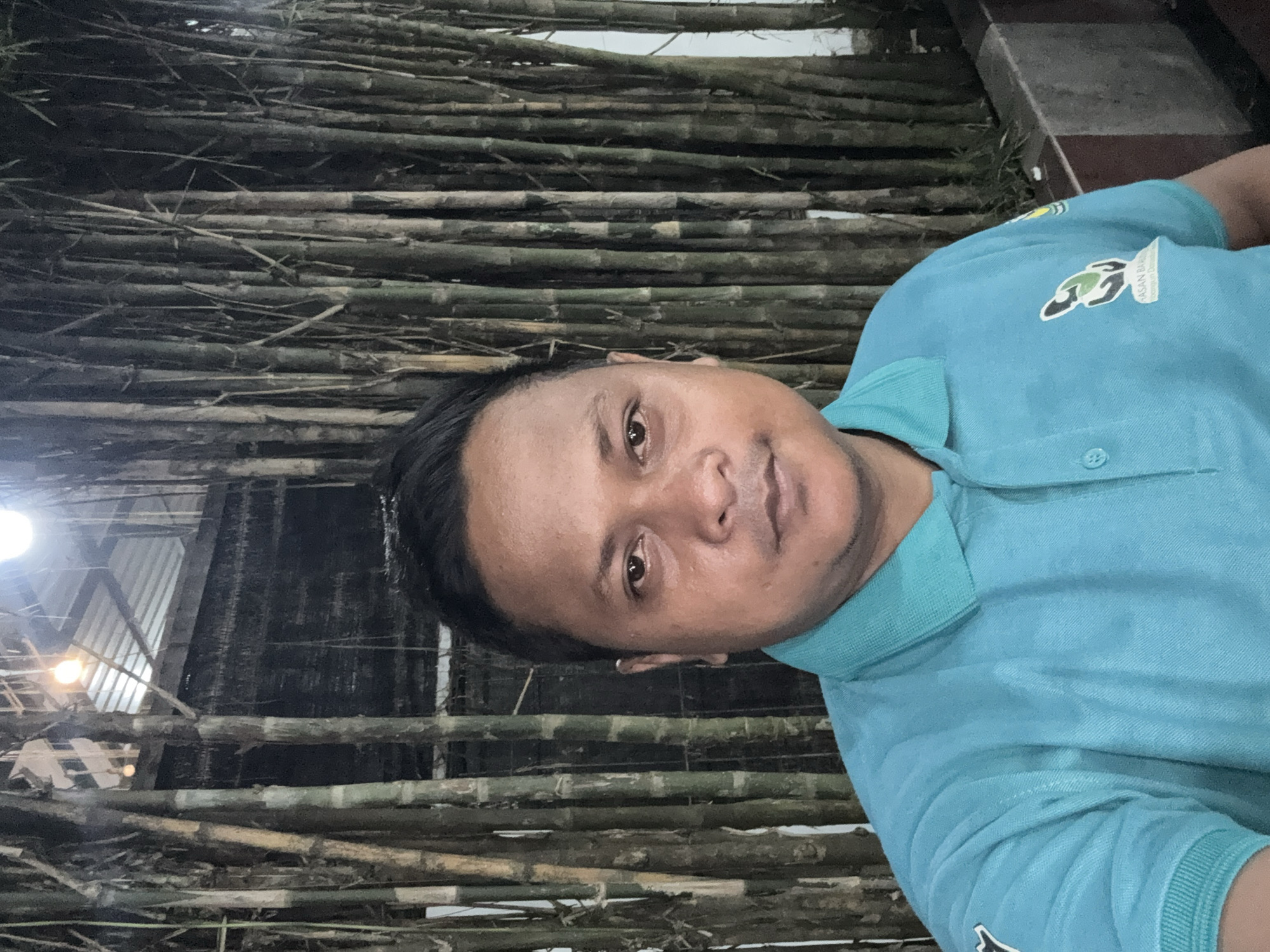 A man is posing for a photo in an outdoor location in Lhokseumawe, Indonesia. The background consists of a densely arranged wall of bamboo stalks, both whole and broken. Parts of a building structure, possibly metal framework, are visible behind the bamboo. The man is wearing a light blue polo shirt with a logo visible on the left chest. The lighting suggests an evening or low-light environment.