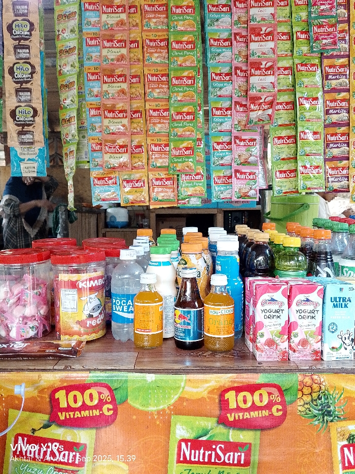The image depicts a retail stall in Sukadana, Indonesia, characterized by a dense display of packaged beverages and snacks. The foreground features a wooden counter laden with various items. On the left are two clear plastic jars with red lids; one contains pink and white candies, and the other, labeled "KIMBO reddi," holds additional snacks. A packaged item lies in front of these jars. To the right, an assortment of bottled drinks is visible, including "POCARI SWEAT," "Red Bull," "YOU-C1000 Orange," and other unidentifiable beverages in clear, yellow, brown, and blue liquid forms. Two pink cartons of "Cimory YOGURT DRINK Strawberry" and one light blue carton of "ULTRA MILK" are also present.
The background is dominated by numerous sachets of instant drink mixes, primarily "NutriSari" in various flavors (e.g., orange, mango, coconut, lime, melon) and "HILO Calcium" chocolate powder, all meticulously hung in vertical rows. Below the hanging sachets, a person with a dark blue head covering and patterned clothing is partially visible, attending the stall with their back mostly turned. Further back, a white and blue container and a green bucket are discernable. A large, colorful banner promoting "100% VITAMIN-C" with "NutriSari" branding and fruit imagery is visible at the very bottom of the frame. The timestamp "Akhlaq & AllSep 2025, 15:39" is imprinted on the lower left.