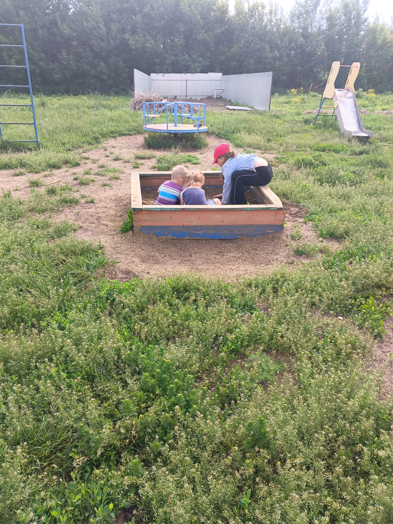 The image depicts an outdoor playground area in Tolbazy, Russia. In the foreground, a sandbox is central to the composition, containing two young children engaged in play. One child, wearing a striped purple and blue shirt, is seated within the sandbox. Another child, also seated, is partially obscured. A third individual, a young person wearing a red cap, denim jacket, and black pants, is kneeling beside the sandbox, seemingly observing or interacting with the children.
The playground itself is situated on a dirt and grassy area. To the left of the sandbox, a blue metal ladder structure, likely part of a larger climbing frame, is visible. Further in the background, a blue metal merry-go-round with red seats is present. To the right of the sandbox, a yellow and light-colored slide stands. A white metal fence runs across the background, with a line of trees beyond it. The ground is uneven, with patches of sand and low-lying green vegetation, some of which appears to be flowering. The overall scene suggests a casual, possibly community-based or residential, outdoor recreational space.
