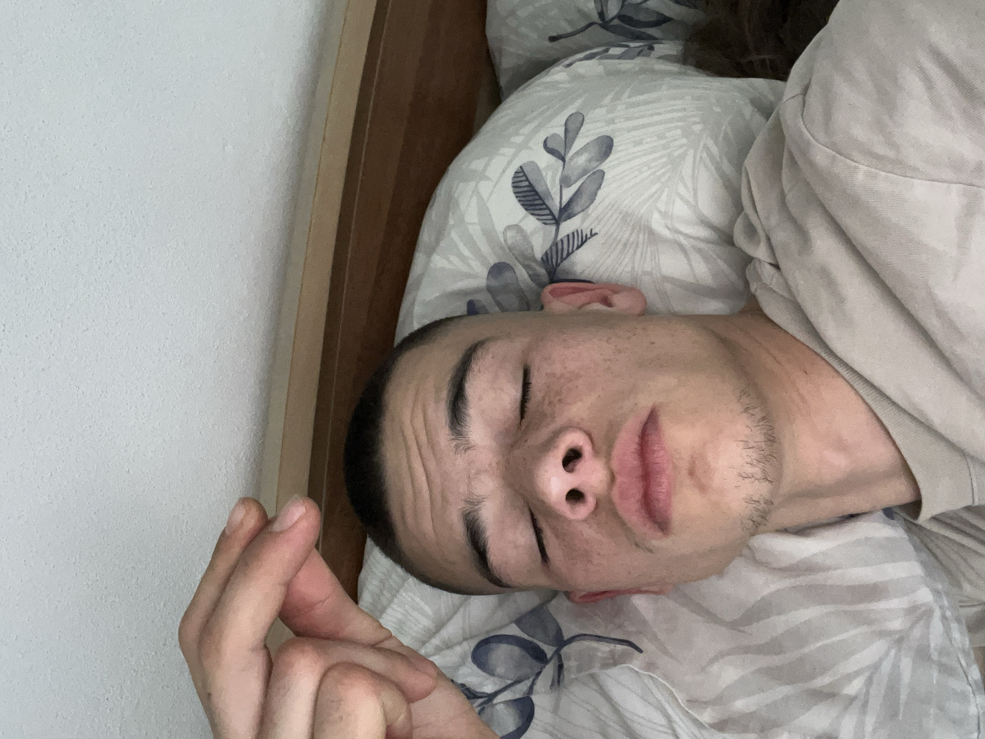A man is lying in a bed. He has short dark hair, closed eyes, and a light complexion with freckles. He appears to be sleeping on a pillow with a white and gray floral pattern. The man is wearing a beige-colored shirt. His hand is raised and partially visible in the lower-left corner of the image. The background includes a white wall and a wooden bed frame. The location is unknown, so the city of Karavelovo, Bulgaria is not discernible in the image.