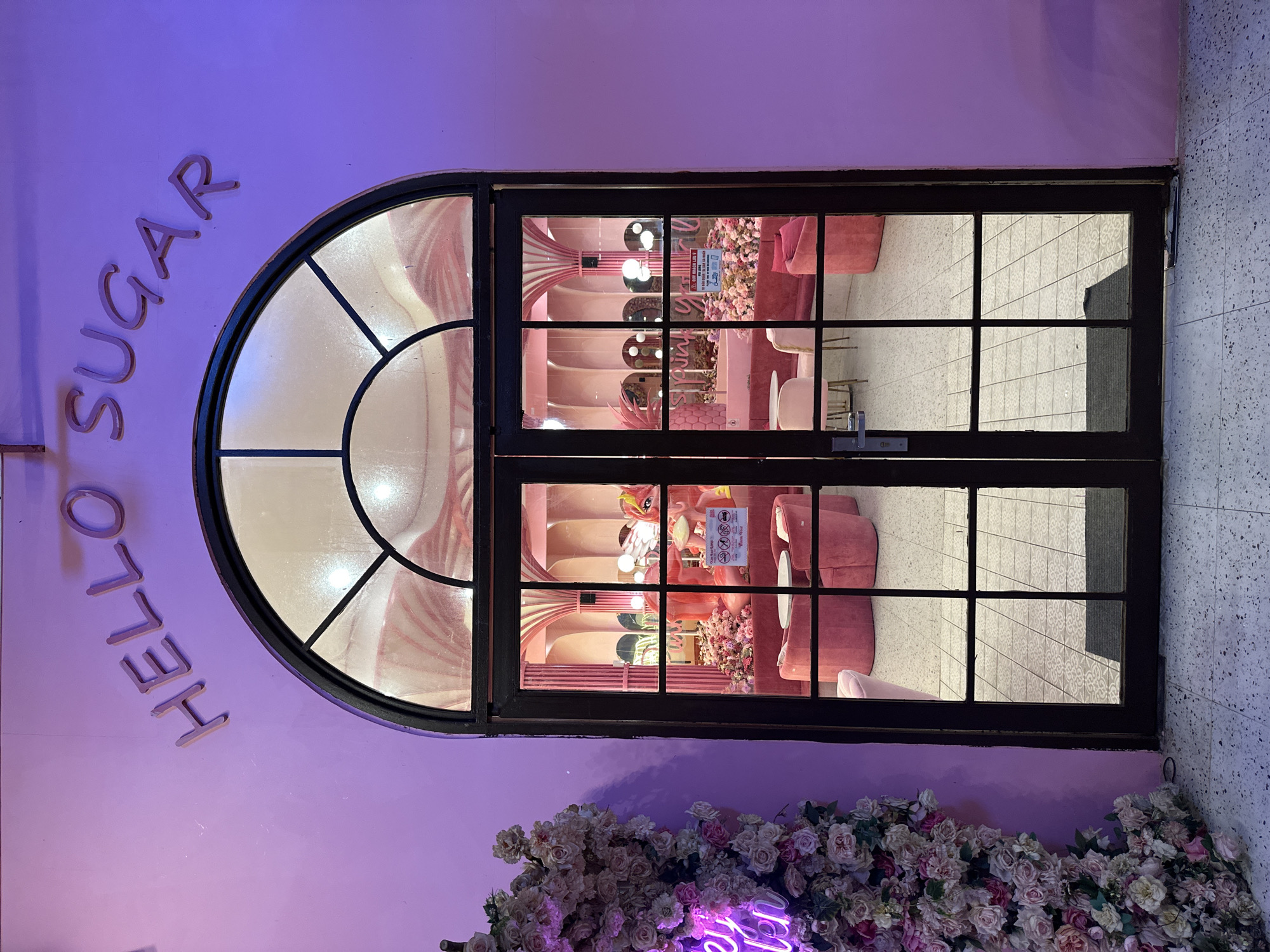The image displays the brightly lit entrance of a vibrant, pink-themed establishment named "HELLO SUGAR" in Tien Thanh Ward, Vietnam, seen at what appears to be evening or night. The exterior wall is a striking purple-pink, adorned with large, light-colored letters spelling "HELLO SUGAR" vertically along the left side.
A prominent feature is the black-framed glass door, which includes an arched top window section and multiple rectangular panes below. Through the glass, the interior of the shop is clearly visible, bathed in warm pink light. The inside features plush pink seating, tables, and ornate decor, including decorative walls with vertical pink stripes, floral arrangements, and what appears to be a flamingo-themed statue or artwork. Several light fixtures cast a soft glow, illuminating the whimsical space. A white sign with red text is visible through the glass panes on the interior back wall, but its content is indistinct.
Along the bottom of the exterior wall, beneath the door, a dense display of artificial flowers in shades of pink, white, and cream creates a lush border. A glowing purple neon sign is partially visible among these flowers, adding to the evening ambiance. The immediate surroundings show a speckled, light-colored wall or pavement on the right, contrasting with the shop's colorful facade. The scene suggests a charming and inviting dessert shop or cafe, designed with a strong, playful aesthetic.