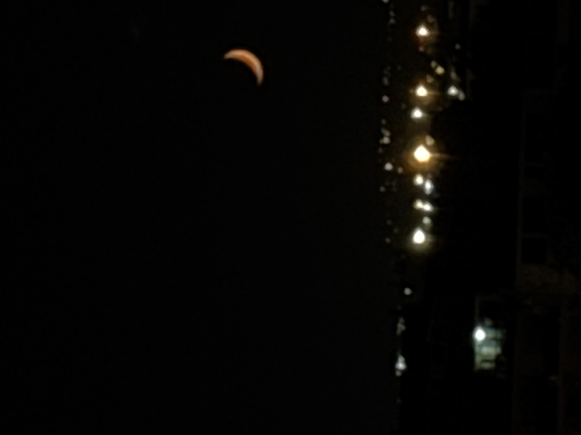 The image shows a crescent moon in the night sky. To the right are out-of-focus lights, appearing to be streetlights, and a building silhouette is visible. The location and city are unable to be verified.