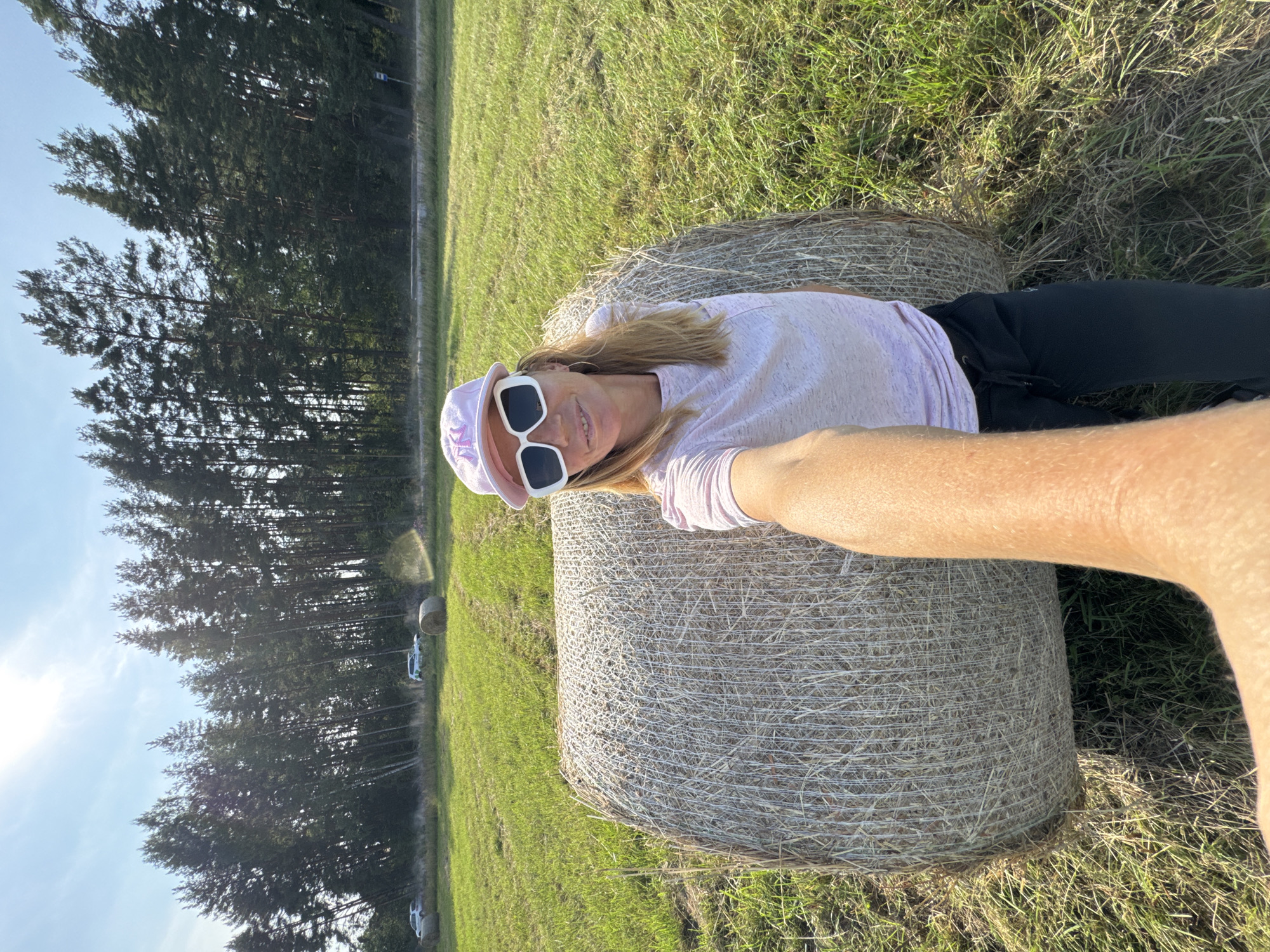 A woman is taking a selfie in a grassy field with hay bales and trees in the background. She is wearing a pink t-shirt, black sweatpants, a pink baseball cap, and white sunglasses. The sun is shining, and there are two cars in the distance. The location is identified as Yablonovka, Russia.