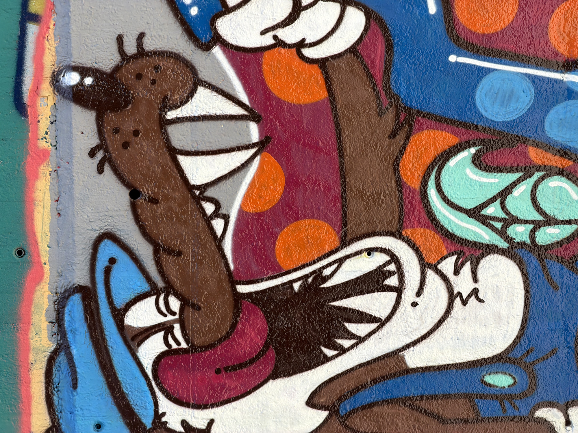 This is a close-up, eye-level, medium shot of a graffiti mural depicting a cartoon wolf character. The wolf has a large, open mouth with sharp, white teeth and a protruding pink tongue. It is wearing a blue hat and appears to have a brown face with white markings around its eyes. The wolf's body is depicted with a burgundy torso covered in orange polka dots. To the right of the wolf's mouth, a white hand is visible, seemingly reaching out. Below the wolf's head, a section of blue is visible, likely part of the wolf's clothing, with a decorative teal element featuring white highlights on its left. The background consists of a textured gray wall with horizontal bands of color at the top: green, yellow, and pink. Two black screw heads are visible in the upper portion of the mural. The graffiti is located on a textured concrete wall, possibly an exterior surface in an urban environment, such as a building facade or a designated graffiti area. This mural is located in the Raval neighborhood of Barcelona, Spain, specifically on a wall adjacent to a parking lot on Carrer de Joaquin Costa.