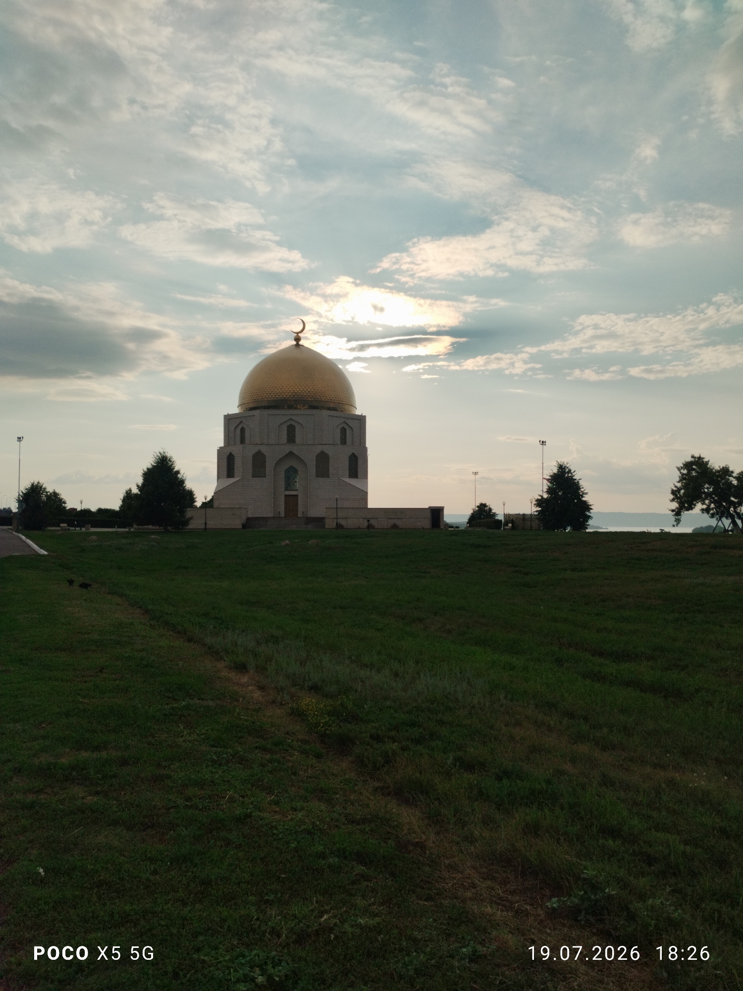 A wide shot captures the white, modern-looking mausoleum of Kurban-Ali Khan in Bolgar, Russia. The structure features a large, golden dome topped with a crescent moon. The mausoleum is situated on a grassy hill, with several trees visible to its left and right. In the background, a body of water stretches towards the horizon, beyond which a hazy landscape is discernible. The sky is filled with dynamic clouds, with sunlight breaking through in the upper right portion of the frame. The bottom of the image contains timestamps and phone model information. There are no people visible in the scene.