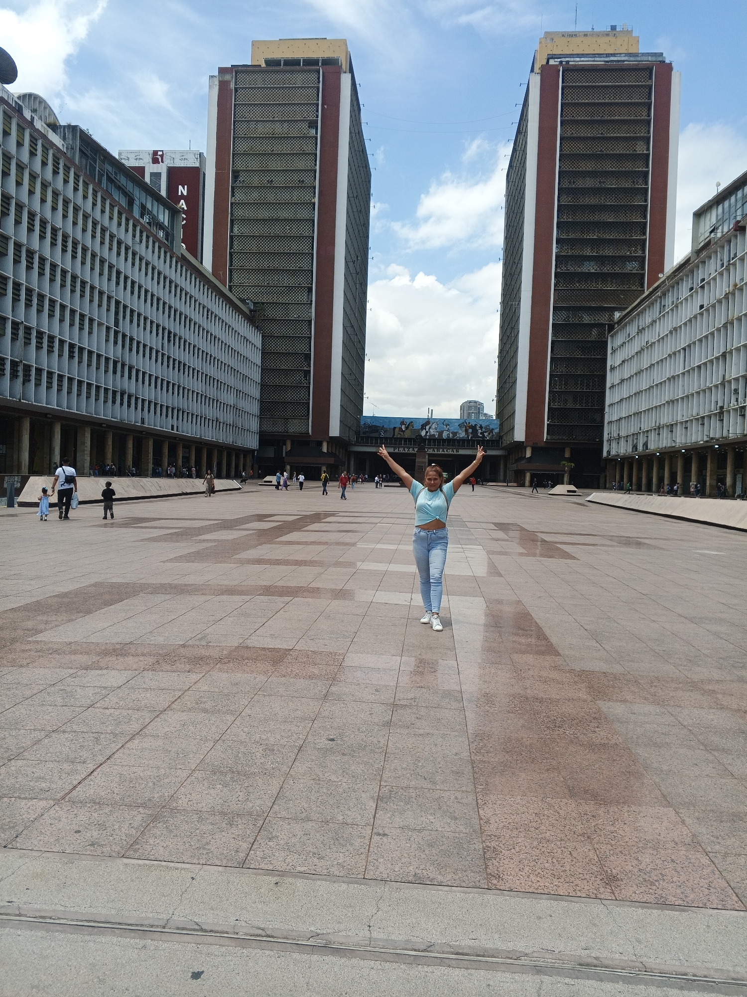 The image depicts a wide, open public square in Caracas, Venezuela, identified by the distinctive architecture of the buildings surrounding it. The square is paved with large, rectangular tiles in shades of gray and pink. A woman stands in the center of the square with her arms raised in a celebratory or expressive gesture. She wears a light blue top, jeans, and white sneakers. In the background, there are two tall, identical buildings with reddish-brown vertical stripes on the sides. These buildings are flanked by lower, elongated structures with repeating patterns of windows and columns. Several other people are scattered throughout the square, including adults and children, some walking alone and others in small groups. The sky above is partially cloudy with patches of blue.