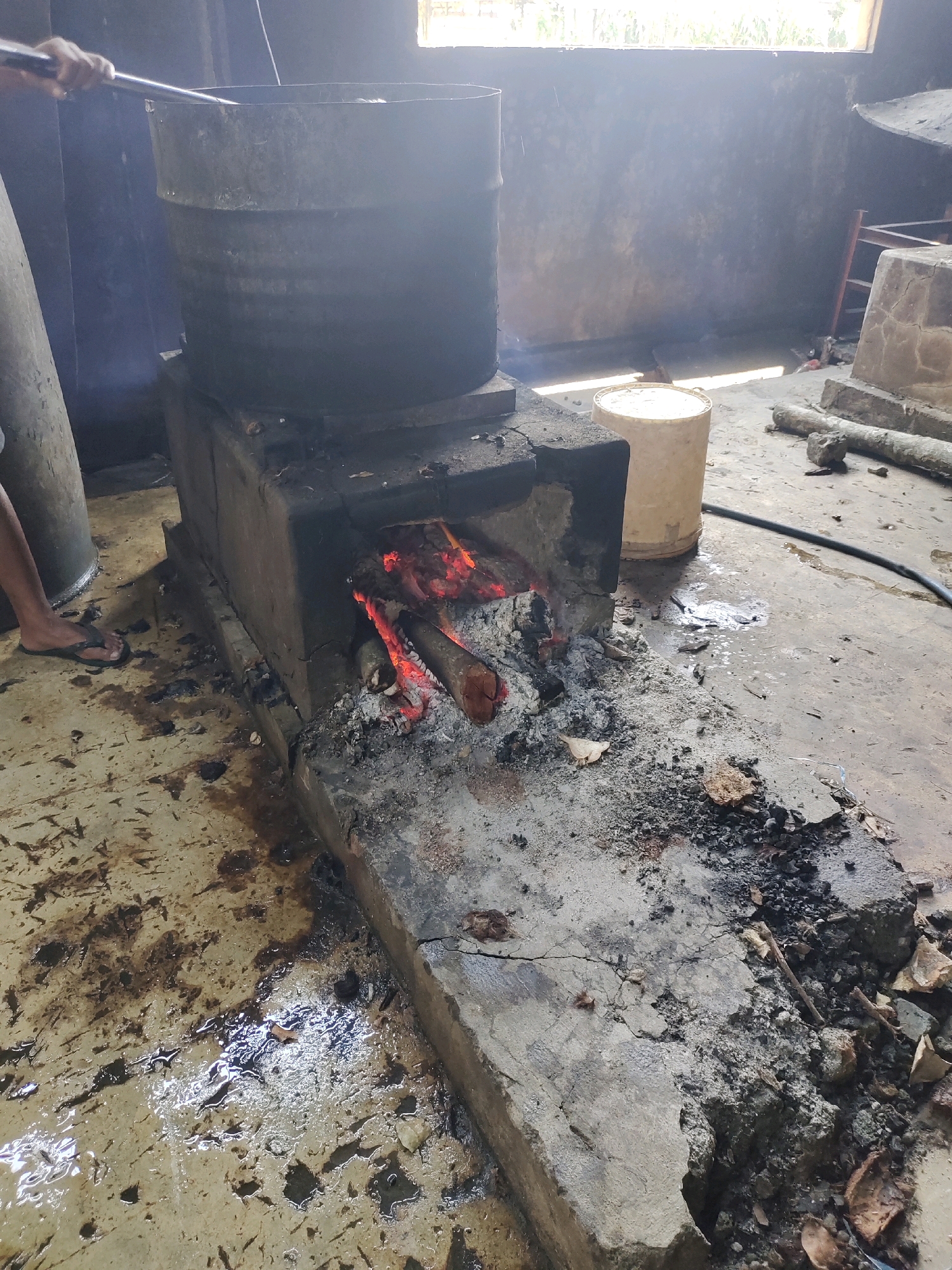 In Ringinrejo, Indonesia, a rustic outdoor cooking setup is captured. A large, dark metal pot sits atop a rough, brick-and-mud stove where a fire burns brightly with visible logs and glowing embers. To the left, a person's leg and foot, clad in a flip-flop, are visible, suggesting they are tending to the cooking. The ground is concrete and appears wet and stained, with debris scattered around the stove, including ash and pieces of what looks like broken concrete or earth. A pale plastic bucket stands to the right of the stove, and a black hose lies on the ground. In the background, a window lets in bright light, illuminating a rough, textured wall. The overall impression is one of a functional, albeit unrefined, outdoor kitchen.