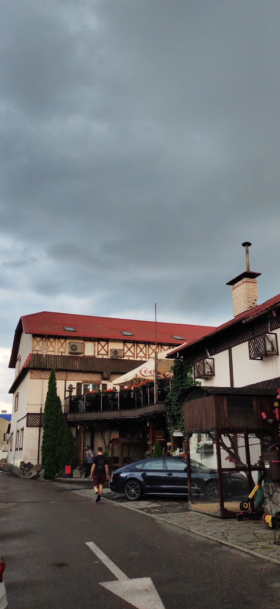 The image depicts an outdoor scene with a building complex under a cloudy sky. The building features a prominent red roof with multiple dormer windows. The facade is a mix of white walls, exposed wooden beams with criss-cross detailing, and dark-trimmed windows. An outdoor seating area with tables and chairs is visible on a balcony-like level, partially covered by a large white umbrella with red accents. A tall, slender evergreen tree stands to the left of the main building. To the right, a smaller, attached structure with a white brick chimney and a dark-trimmed roof is visible. In the foreground, a dark-colored sedan is parked on the asphalt. A person wearing a dark t-shirt and burgundy shorts is walking away from the camera towards the parked car. The ground is wet, suggesting recent rain. The scene appears to be a restaurant or similar establishment. While the image is attributed to Homyel, Belarus, no specific landmarks or identifying features within the image confirm this location.