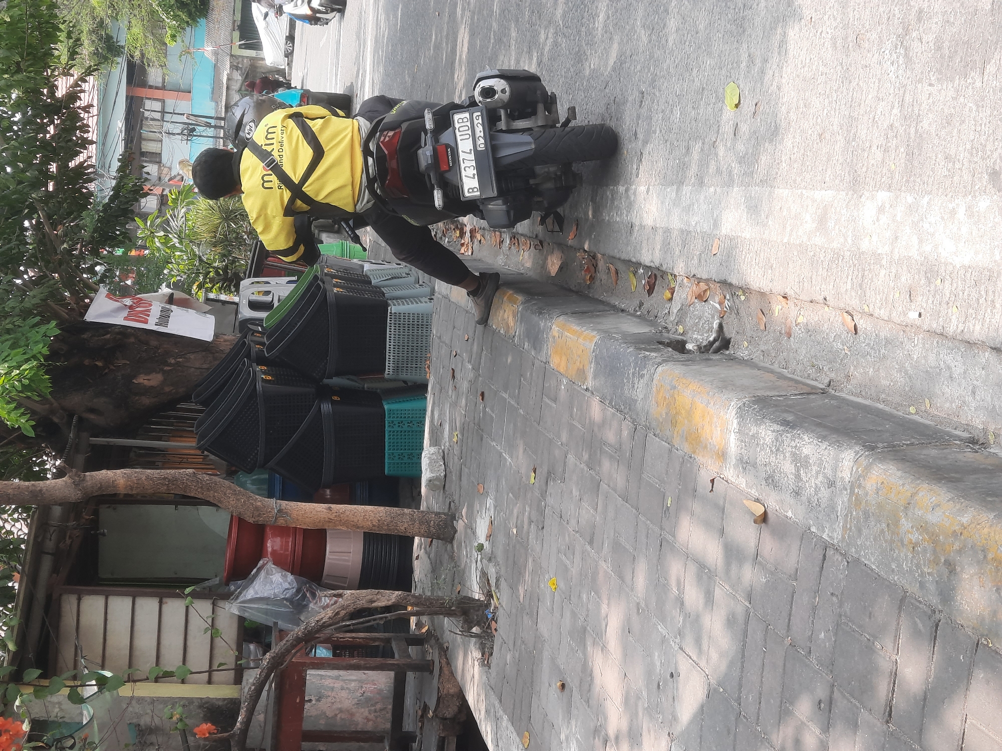 A male individual wearing a yellow Maxim ride-sharing jacket is seated on a dark-colored motorcycle parked along a sidewalk in North Jakarta, Indonesia. The motorcycle's license plate reads "B 4374 UDB". The sidewalk is paved with gray bricks and bordered by a curb with yellow markings. Behind the motorcycle, a stack of black baskets and red and cream-colored plastic containers are visible near a small roadside structure. Trees with green foliage are present in the background. The scene depicts a street setting with typical urban elements.