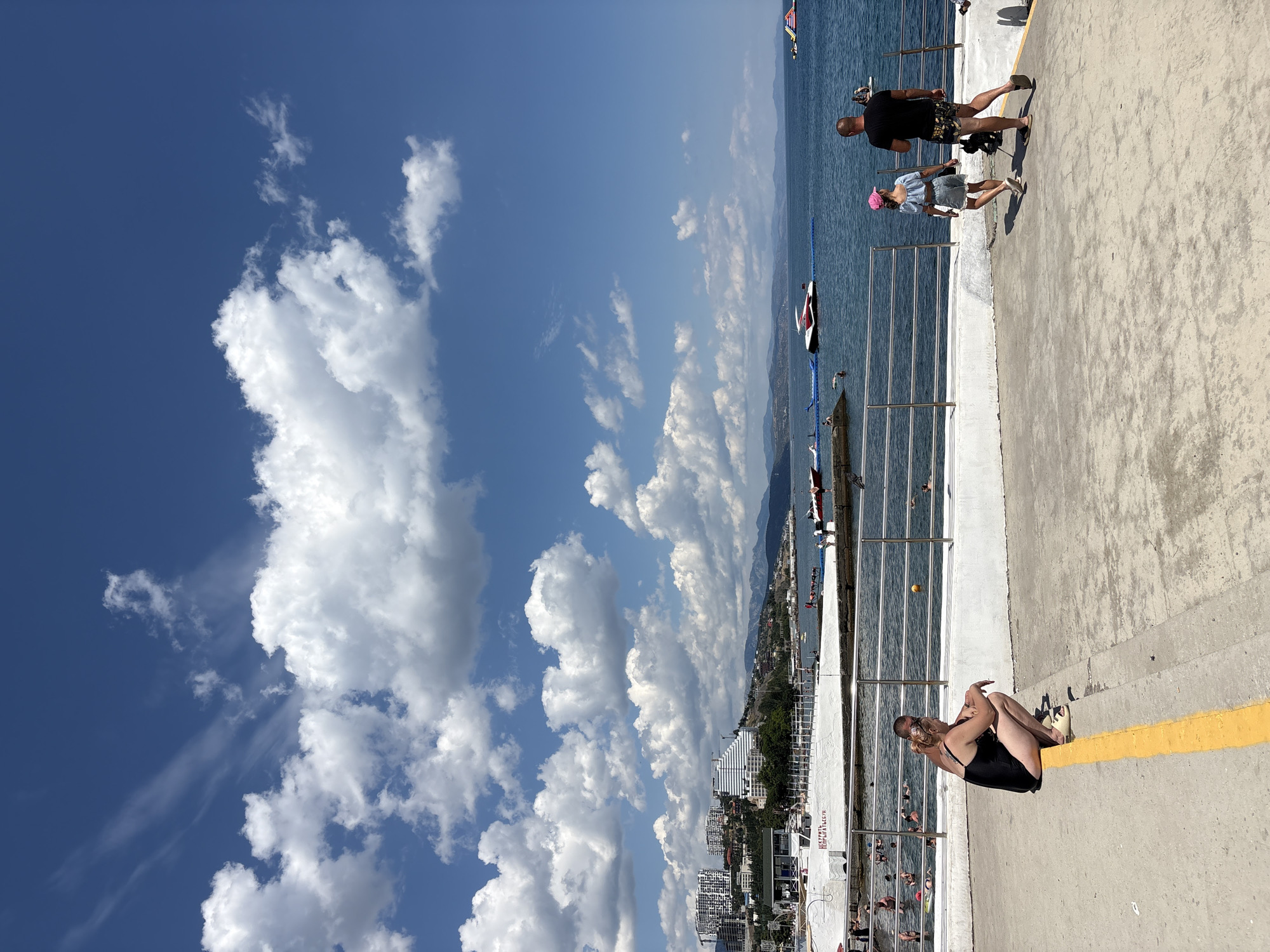 The image depicts a sunny day at a beach in Alushta, Ukraine. In the foreground, a woman in a black swimsuit is squatting on a concrete walkway, facing the sea. To her right, a couple walks away from the camera, with the man wearing a black t-shirt and patterned shorts, and the woman wearing a light blue top, shorts, and a pink hat. A small dog walks beside them.
Beyond the walkway, there is a metal railing separating the promenade from a beach area. People are seen swimming and relaxing in the water, and several boats are moored in the sea. In the background, the city of Alushta is visible with its buildings and mountains. The sky is blue with scattered white clouds. The overall scene suggests a typical summer day at the seaside.
