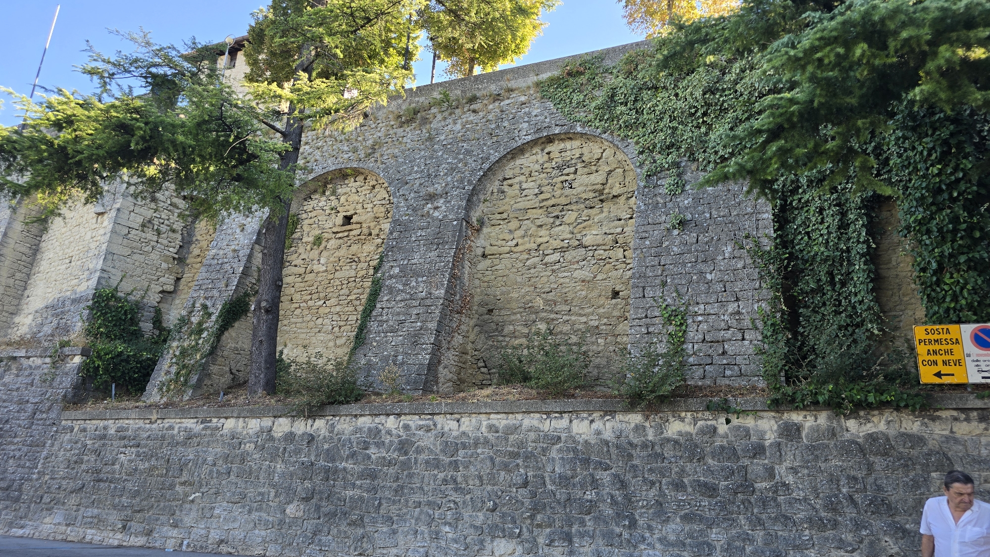 A man in a white polo shirt stands on the right side of the frame, looking downwards. He is positioned in front of a stone wall that forms the foreground of the image. This wall is constructed from large, roughly hewn grey stones. Above this wall, a section of a much larger, ancient stone fortification rises. This upper wall is built from yellowish stones and features several large arched openings that are filled in with rubble and masonry. The fortification is overgrown with ivy and other greenery, and a large tree grows from the middle section, its branches extending over the wall. The sky above is a clear, bright blue. To the far right of the upper wall, a yellow sign with black text and arrows is visible.