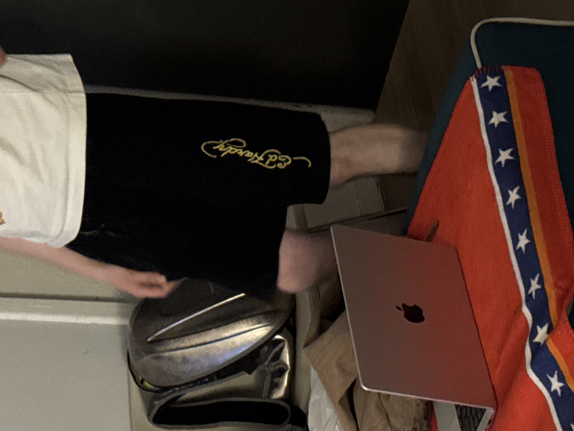 A person stands indoors, wearing a white t-shirt and black shorts with a gold "Ed Hardy" logo. In the foreground, a silver laptop sits open on a red blanket patterned with stars and stripes, resembling a Confederate flag. To the left, a metallic, rounded backpack rests against a wall. The background shows a plain wall and a dark doorway. The scene suggests a casual, indoor setting.