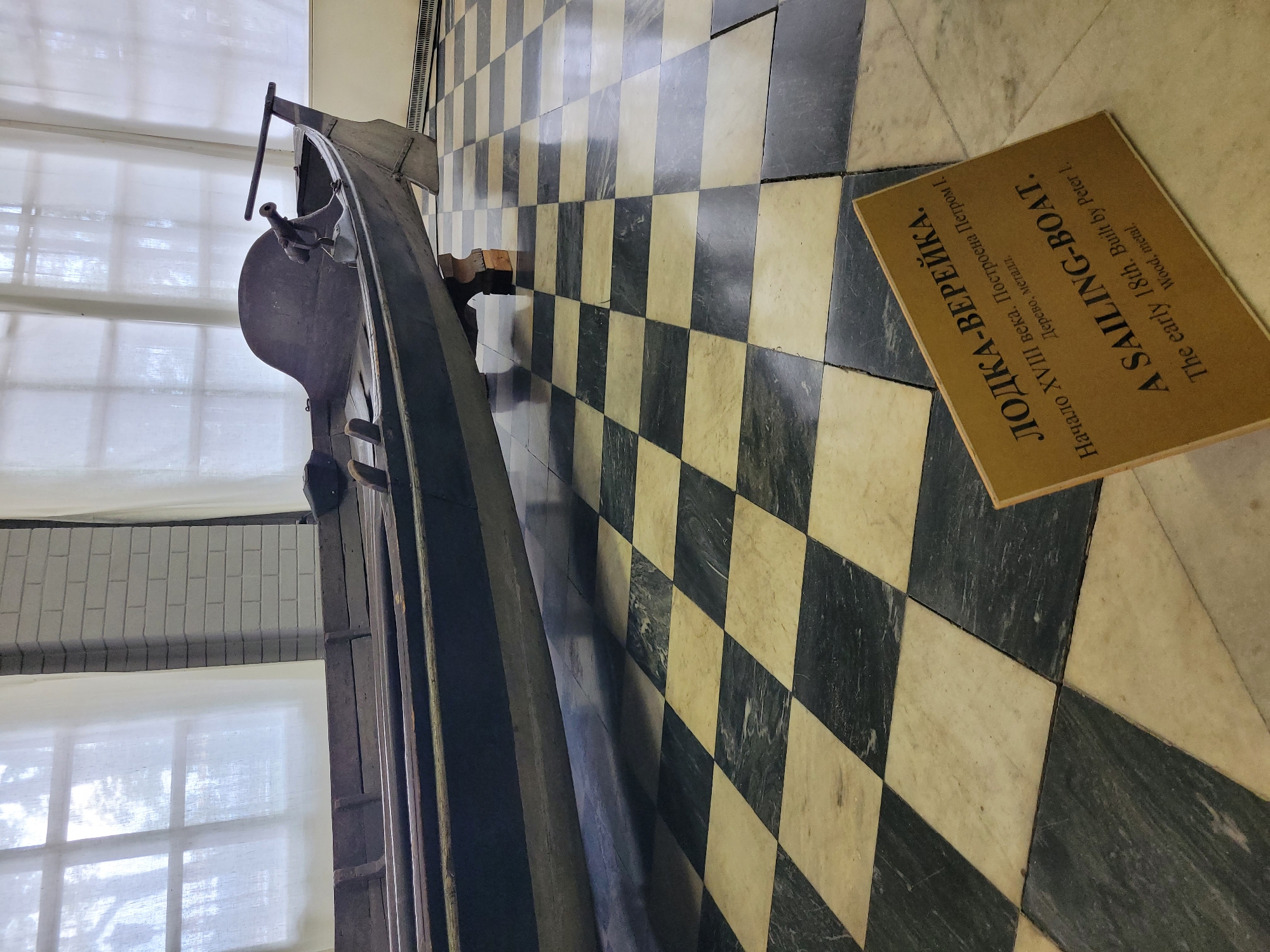 The image shows an antique wooden boat on display in a museum. The boat is dark in color and appears to be a sailing boat, as indicated by the inscription on a sign in front of it. The sign is made of cardboard and has text in both Russian and English. The Russian text reads "ЛОДКА-ВЕРЕЙКА. Начало XVIII века. Построена Петром I. (Дерево, металл.)" and the English text reads "A SAILING-BOAT. The early 18th. Built by Peter I. (Wood, metal)". The boat is supported by wooden blocks and rests on a black and white checkered floor, likely made of marble. In the background, there are large windows covered with sheer white curtains, and a brick wall. The lighting in the room is soft and diffused, suggesting it's either overcast outside or the lighting is intentionally subdued for display purposes. The boat is positioned at an angle, with its stern facing the viewer and its bow extending to the left. A small detail on the stern appears to be a decorative element or part of the steering mechanism. The environment appears to be indoors, likely a museum or historical exhibition space, given the display of the artifact and the informational sign. The overall impression is one of historical preservation and display in a controlled environment.