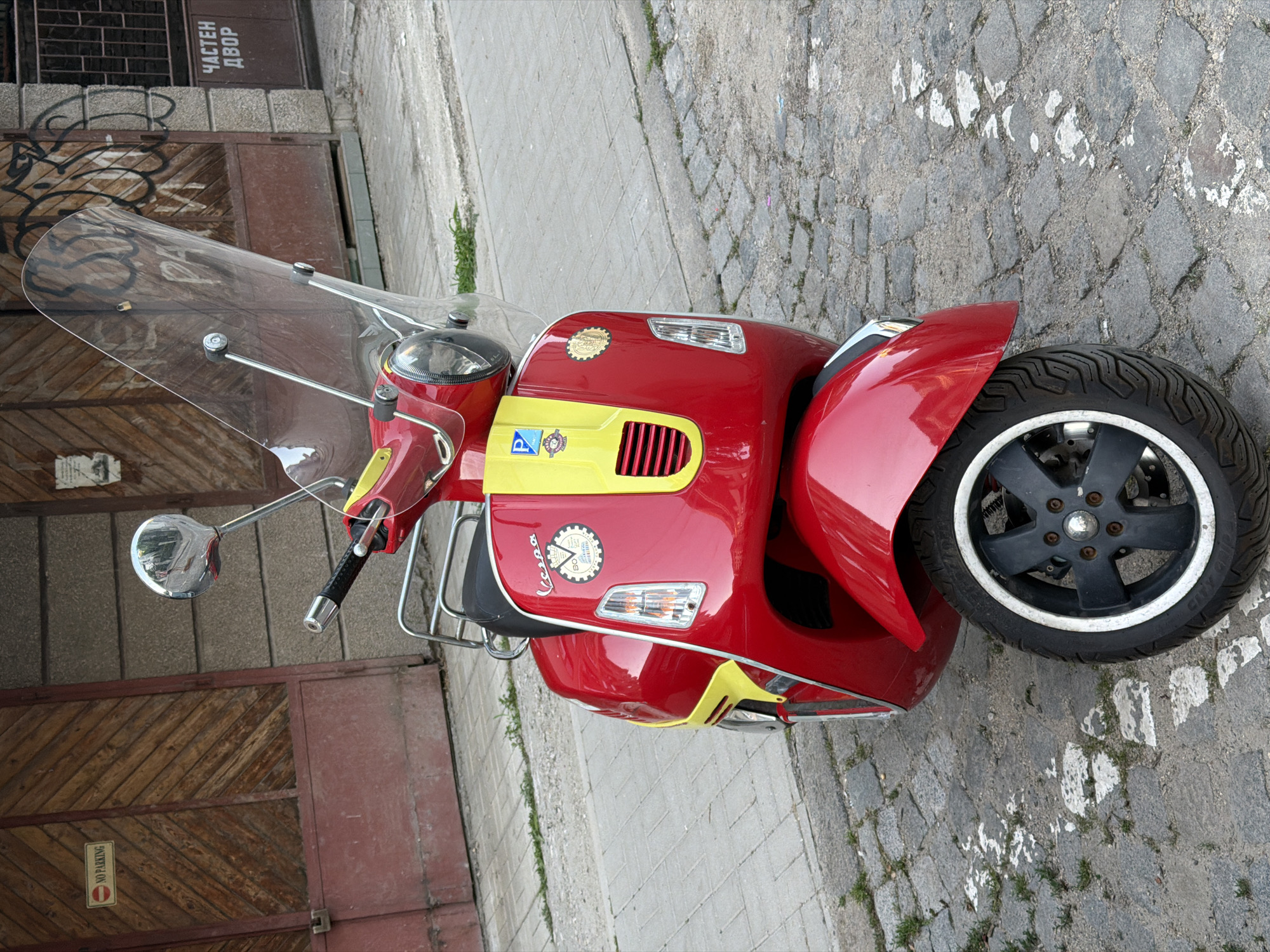 A red and yellow Vespa scooter is parked on a cobblestone street in front of a building with a wooden door. A "No Parking" sign is visible on the door. Graffiti is spray-painted on another section of the building's facade. A sign on the building reads "Частен Двор" (Private Yard). The scooter has a clear windshield and a metal luggage rack. The street is lined with some sparse green vegetation. The scene appears to be in an urban environment.