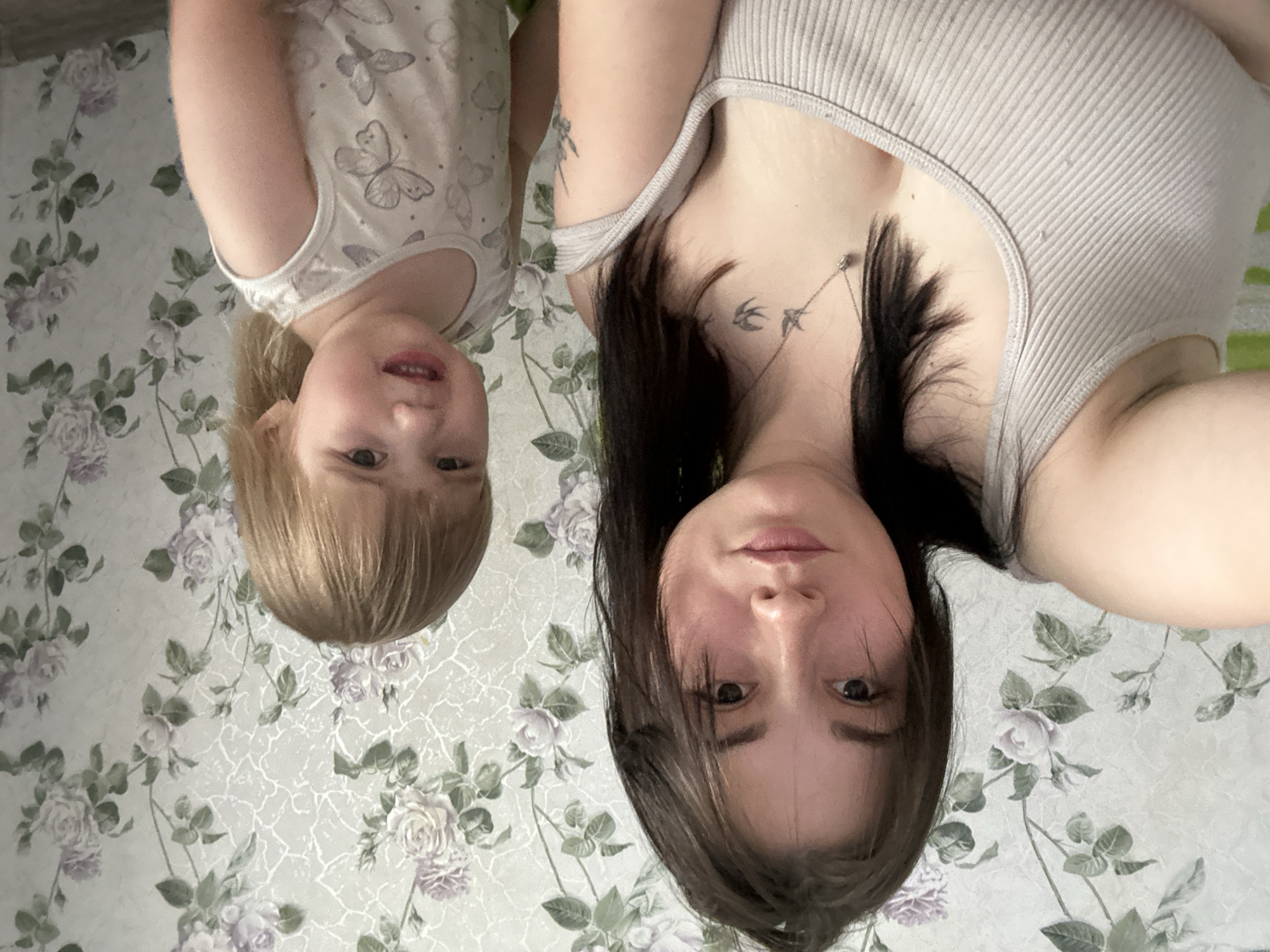 A young woman with dark hair and a light-colored tank top is taking a selfie with a young child. The child, wearing a light-colored shirt with butterfly patterns, is looking towards the camera with a slightly distressed expression. They are standing in front of floral wallpaper. The setting appears to be indoors, possibly a home environment in the city of Chaikovsky, Russia. The woman has tattoos visible on her chest and arm.