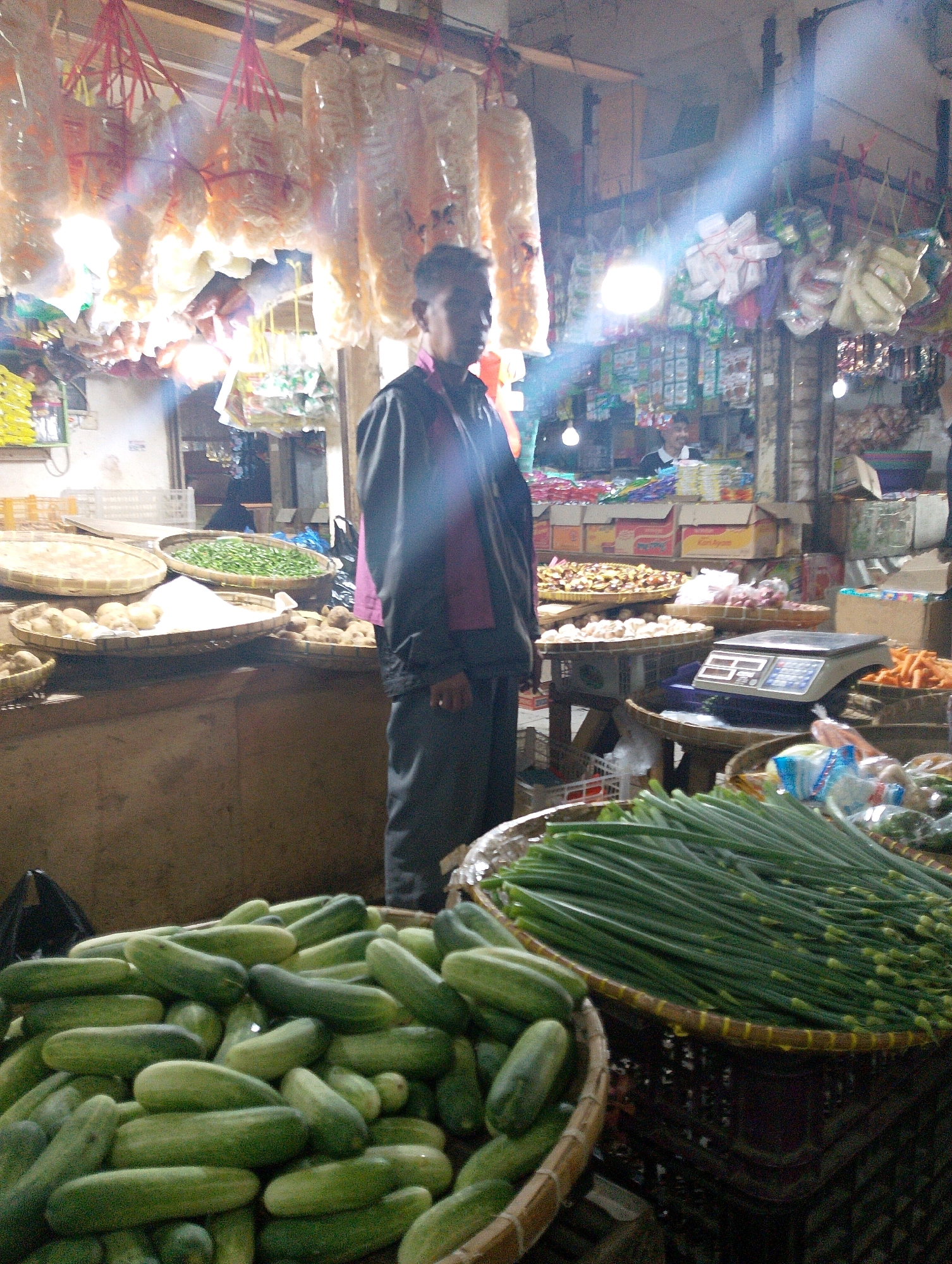 The image shows a market stall in Lembang, Indonesia, with a vendor standing behind a display of fresh produce. The vendor, a man with dark hair, is wearing a dark jacket over a pink shirt and dark pants. He is looking towards the left side of the frame, with his right hand resting on his waist. In front of him, on a wooden counter, are various food items displayed in woven baskets. These include piles of cucumbers in the foreground, green onions, potatoes, and what appear to be green beans or peas. Behind the counter, shelves are stocked with packaged goods, including boxes of instant noodles and other food items. Hanging from the ceiling are various types of dried goods, possibly snacks or crackers, packaged in translucent plastic. The market appears to be indoors, with artificial lighting contributing to the scene's illumination, and a noticeable light flare originating from the upper right. The overall environment suggests a bustling local market.