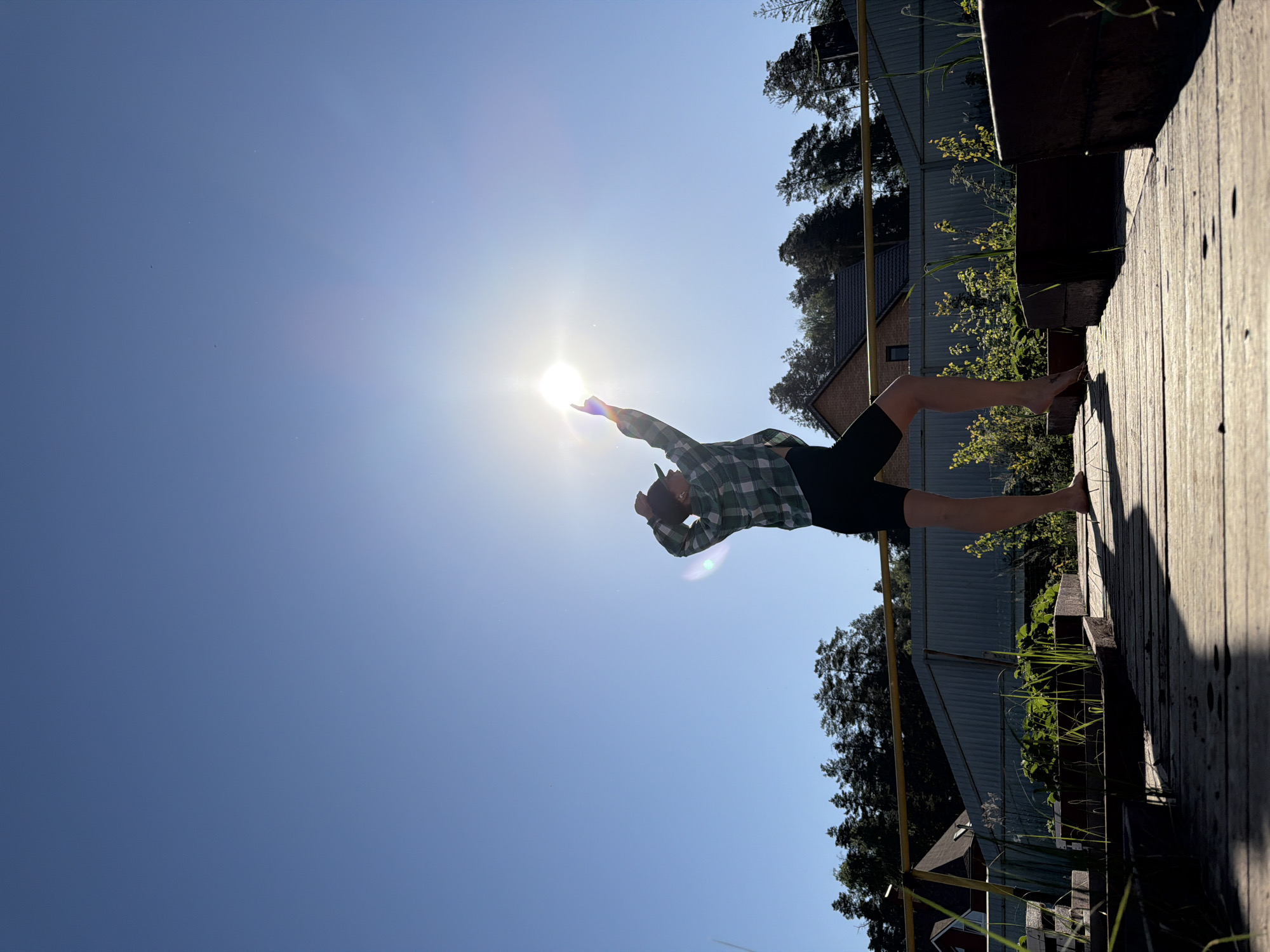 A man stands barefoot on a wooden walkway with his arm extended towards the bright sun. The man is wearing a plaid shirt and dark shorts. The walkway is elevated and overlooks a residential area with houses and trees. The sky is clear and blue. The location appears to be in Chaikovsky, Russia based on the landscape and architecture visible.