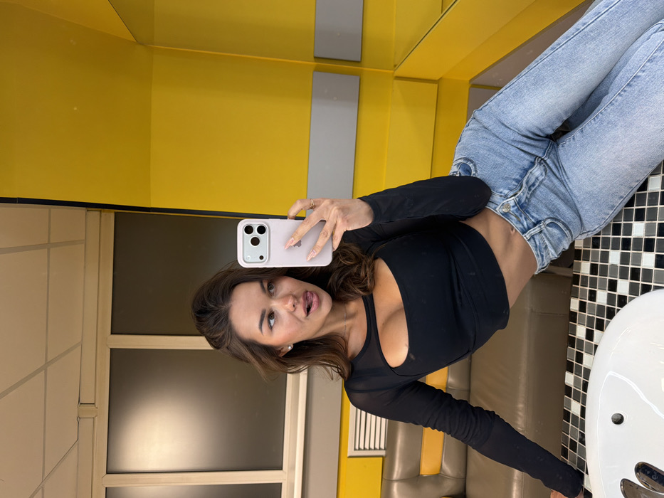 A single female subject is depicted taking a mirror selfie in what appears to be a restroom or lounge area within Tselinny Cafe (Целинный) at Mira Avenue, 86, Krasnoyarsk, Russia. The woman, appearing to be in her 20s or early 30s, has long brown hair and is wearing a black long-sleeved crop top with a chest cut-out and light blue denim jeans. She has small stud earrings and a ring on her right hand. Her expression is playful, with her tongue slightly extended from her mouth. She holds a light pink/purple cased iPhone in her right hand, angled towards an unseen mirror.
In the immediate foreground, a white sink basin and a countertop with a black and white checkered tile pattern are visible. To the left of the subject, behind her, is a segment of a brown leatherette booth featuring a horizontal yellow stripe along its backrest. Above the booth, a dark-tinted rectangular window or glass partition with a light-colored frame is present, with a ventilation grille located below it. The background on the right is dominated by bright yellow wall paneling, which is also reflected in a mirror positioned to the right. The ceiling is light-colored with visible grid lines.