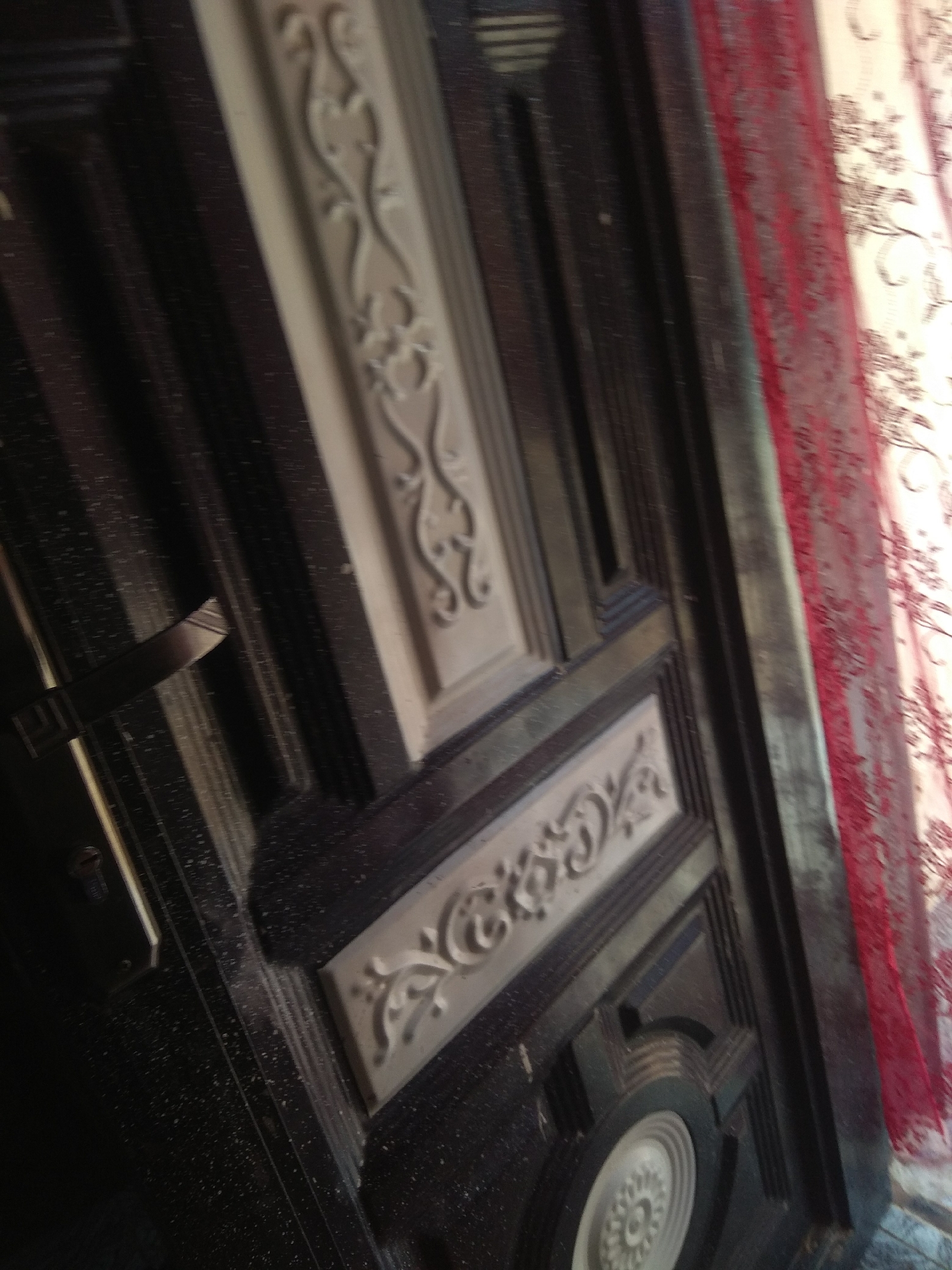 The image shows a close-up, angled view of a dark-colored, ornate door. The door features raised decorative elements in a lighter color, with intricate floral or vine-like patterns. A metallic door handle is visible on the left side of the frame, extending outwards. The background on the right side of the image shows a red, sheer curtain with a patterned design. The scene appears to be indoors or in a sheltered area, with dust particles visible on the door's surface. The context of Jalingo, Nigeria is not evident from the visual information provided in the image.