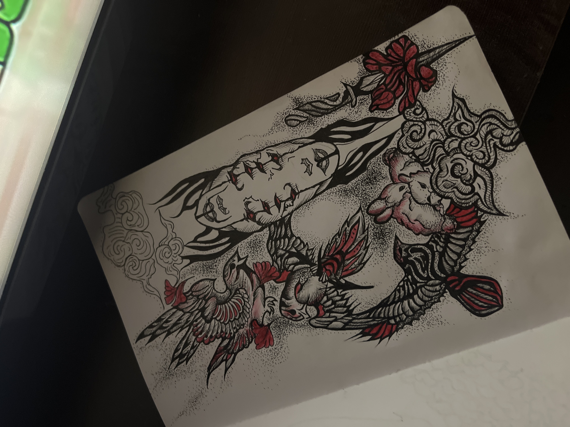 The image captures an intricate, stylized drawing on a white sheet of paper, presented from an overhead, angled perspective. The artwork is executed primarily in black ink with striking red accents, characterized by detailed linework and extensive use of stippling for shading and texture.
The central subjects of the drawing are several stylized birds, possibly mythical creatures like phoenixes, depicted in dynamic flight or action. Their wings and bodies are rendered with elaborate black patterns, while elements such as their plumage and facial features are highlighted in vibrant red.
Extending diagonally from the upper left, a long, dark, stylized object resembles a weapon or organic form, featuring distinct red "stitches" or markings along its length, and subtle, integrated eye-like shapes. Adjacent to this object, at the top right of the drawing, are detailed red floral elements with associated leaves.
The composition also incorporates swirling, cloud-like patterns, consistent with traditional Asian art motifs, which frame and intersperse with the main figures. A soft, pinkish, creature-like form, possibly a stylized animal head or part of a larger being, is partially visible within these cloud elements on the right.
The white paper lies on a dark, flat surface, likely a desk or table. A portion of a digital screen, displaying indistinct green and light-colored graphics, is visible on the far left, suggesting an indoor, possibly artistic, workspace. There is no discernible text within the image. The style evokes a modern illustrative or neo-traditional tattoo aesthetic.
