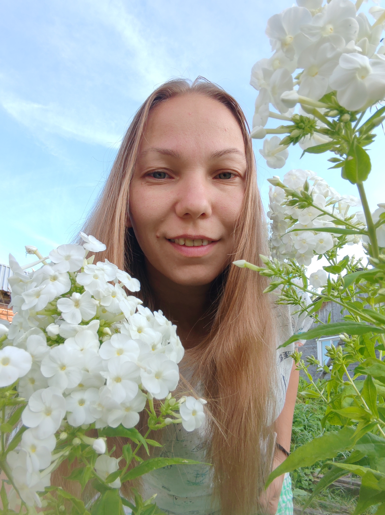 A young woman with long, blonde hair is photographed from a low angle, with white flowers in the foreground partially obscuring her face. She is smiling and looking directly at the camera. The background shows a blue sky with wispy clouds and greenery, suggesting an outdoor setting, possibly a garden or yard. The image appears to have been taken in the city of Chukhloma, Russia.