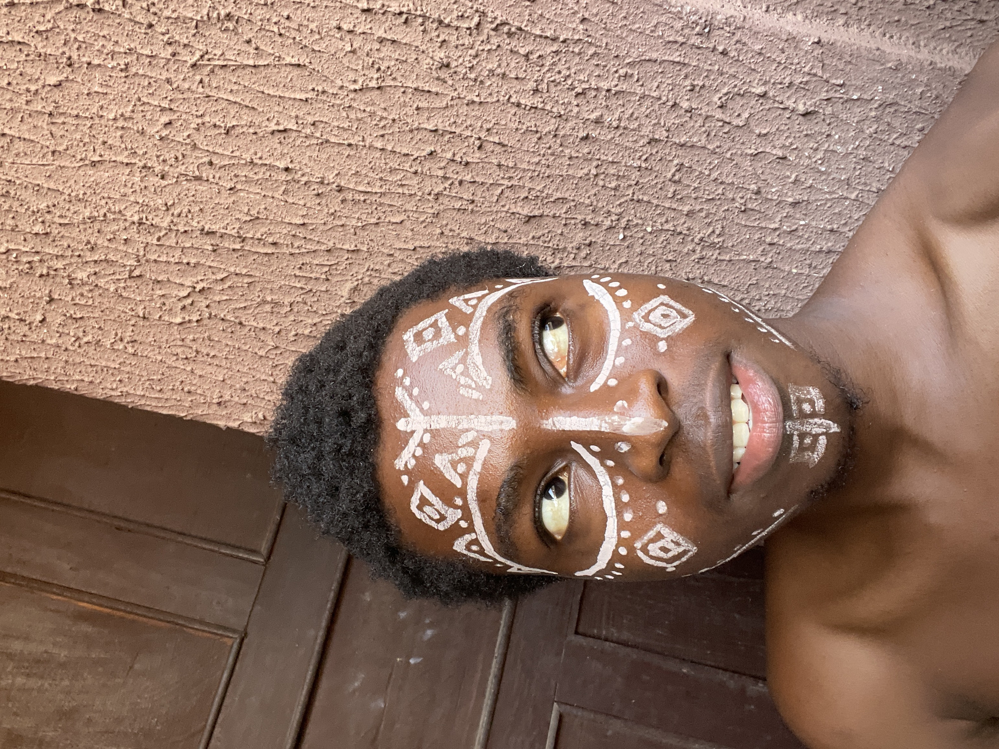 A close-up photograph of a shirtless Black man with short, dark hair and white painted tribal designs on his face. He is lying on a dark wooden surface or floor with his head angled towards a textured beige wall. He is looking at the camera with his mouth slightly open, revealing his teeth. The location is unspecified and Nkpor, Nigeria is not verified in the scene.