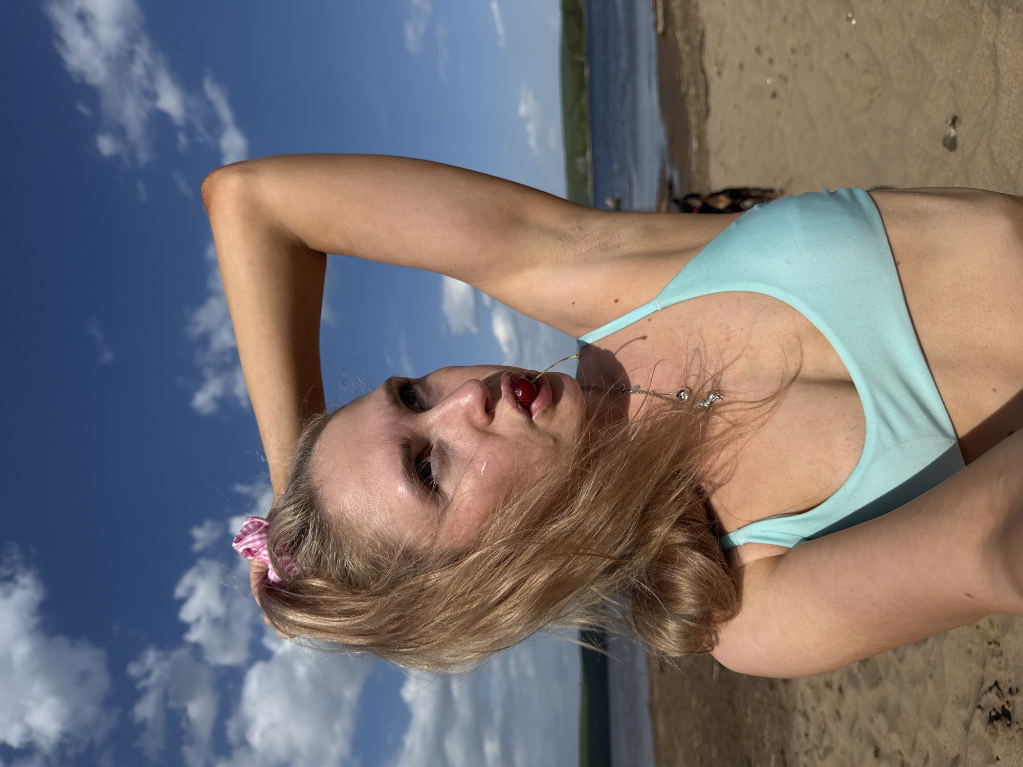 A woman is lying on a sandy beach with her head tilted back. She has fair skin, blonde hair, and is wearing a light blue bikini top. A cherry is positioned in her mouth. Her left arm is raised and bent, with her hand near her head, and her right arm is partially visible in the lower right corner of the image. A pink and white checkered scrunchie is in her hair. A silver necklace adorns her neck.
The background shows a body of water with trees visible on the opposite shore. The sky is blue with scattered white clouds. The sandy beach has some debris or markings.
Based on the prompt, this scene is located in the city of Chaikovsky, Russia.