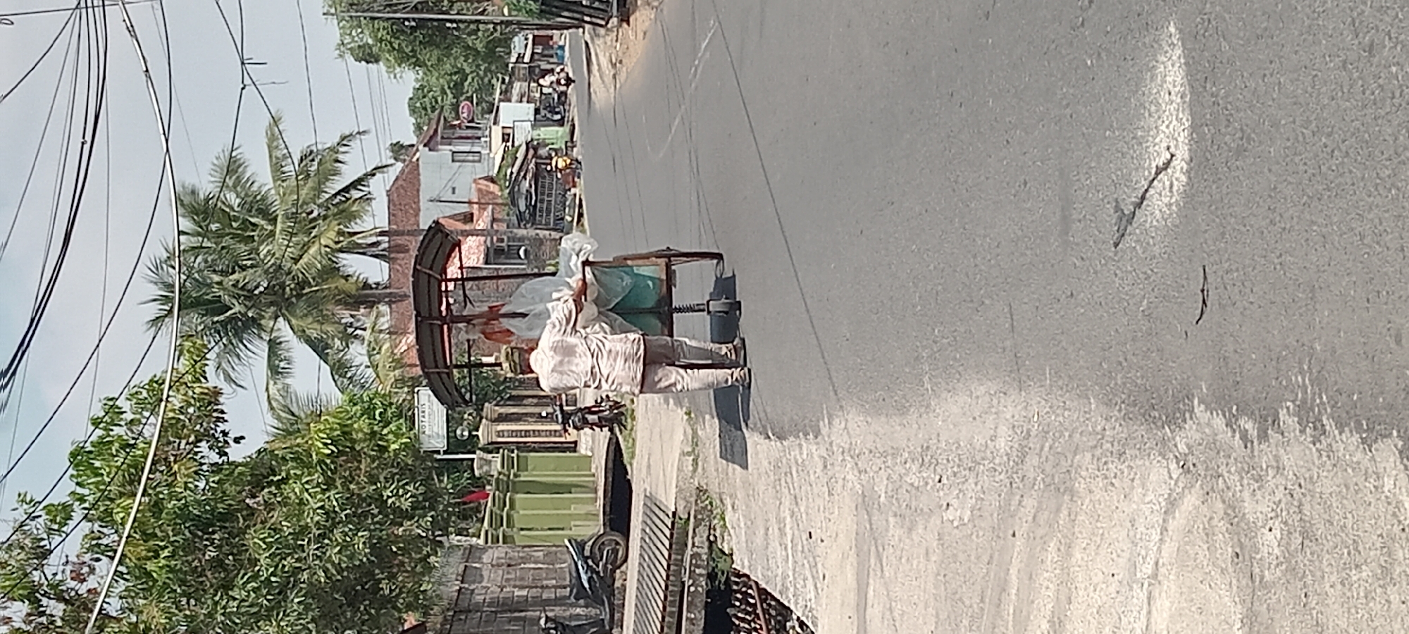 In Wanasigra, Indonesia, a man is pushing a cart along a paved road. He is wearing a light-colored, long-sleeved shirt and pants, and a patterned cap. The cart has a metal frame and a fabric covering, and it appears to be carrying white material, possibly goods for sale or delivery. The road is bordered by a concrete sidewalk on the left, and a small drain or ditch runs alongside the sidewalk. The background shows houses with tile roofs, trees, and power lines overhead. A motorcycle is parked on the left side of the road near a fence. The sky is clear and bright, suggesting a sunny day.