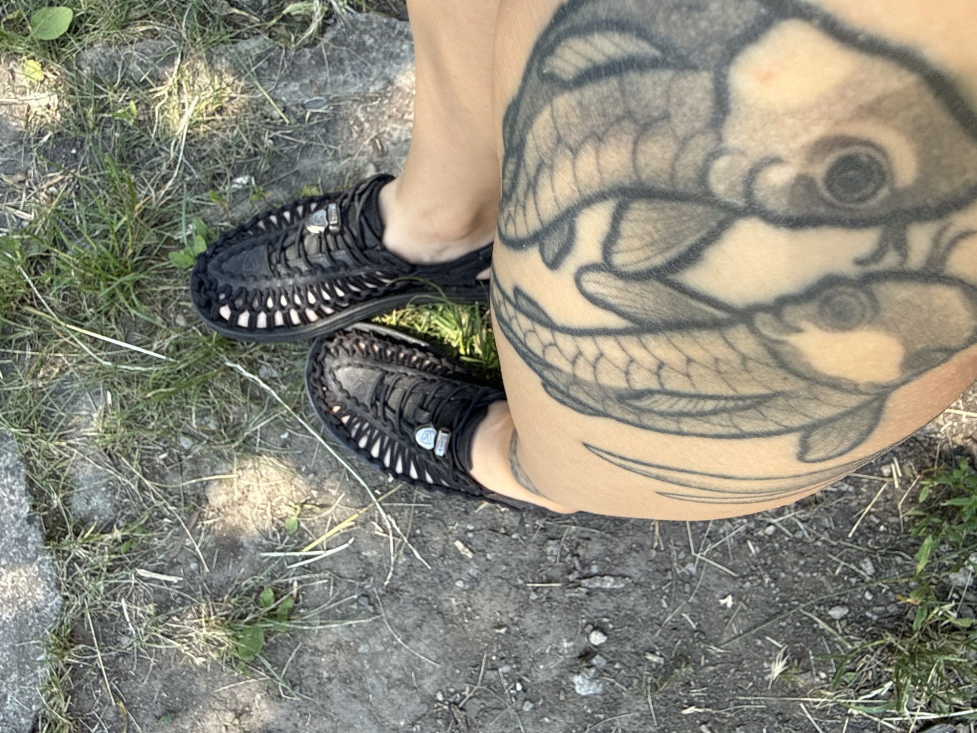 The image shows a person's legs and feet from a high angle, with a tattoo on the thigh. The person is wearing black sandals with white lacing. The ground is covered in dirt and grass. The scene appears to be outdoors.