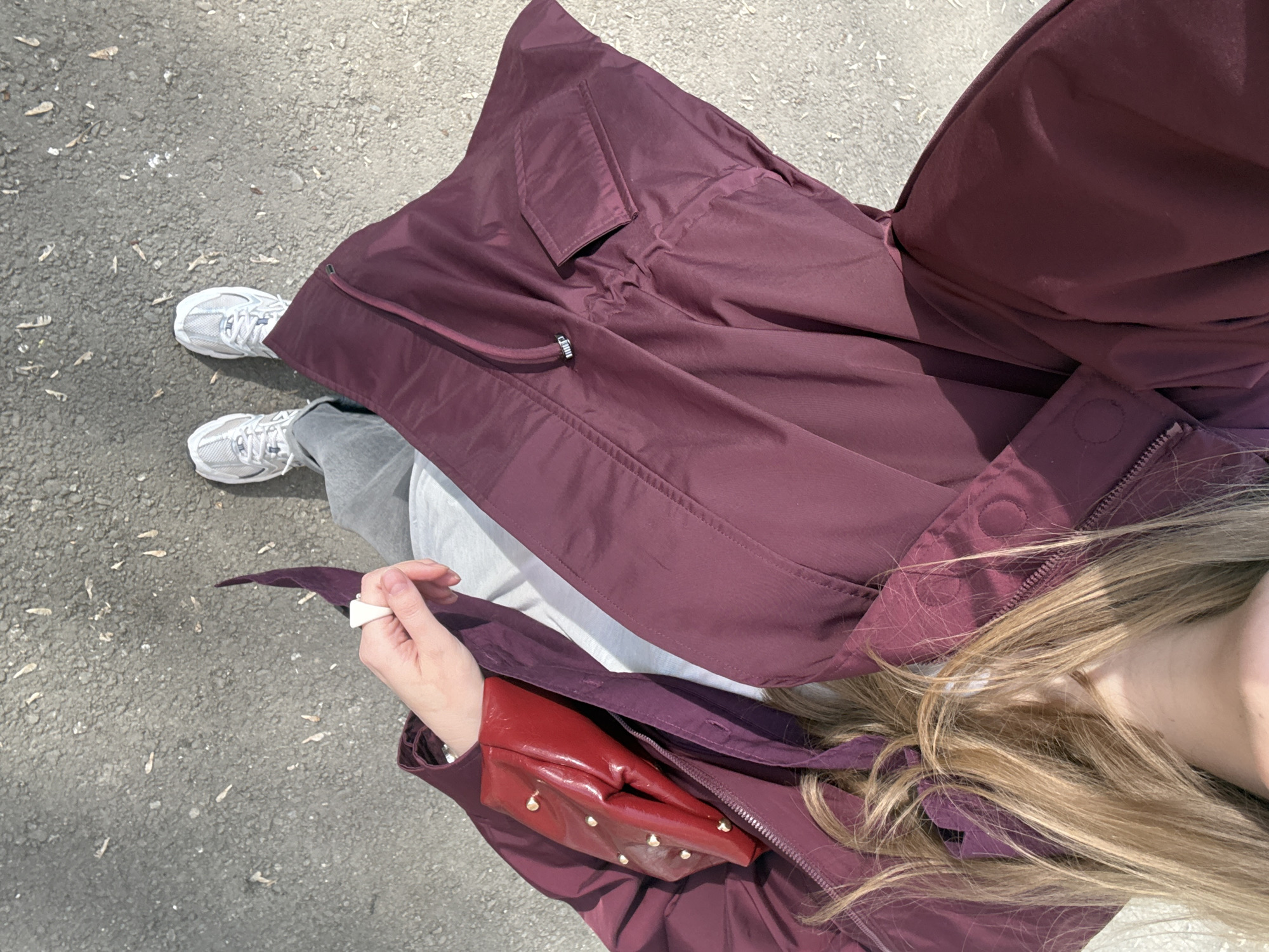 A person, seen from a high-angle perspective, stands on a paved surface. The person wears grey sneakers, grey pants, and a large, open, burgundy-colored jacket. The person also carries a small, red handbag with gold embellishments. The background is a grey, paved surface. The location cannot be determined from this image.
