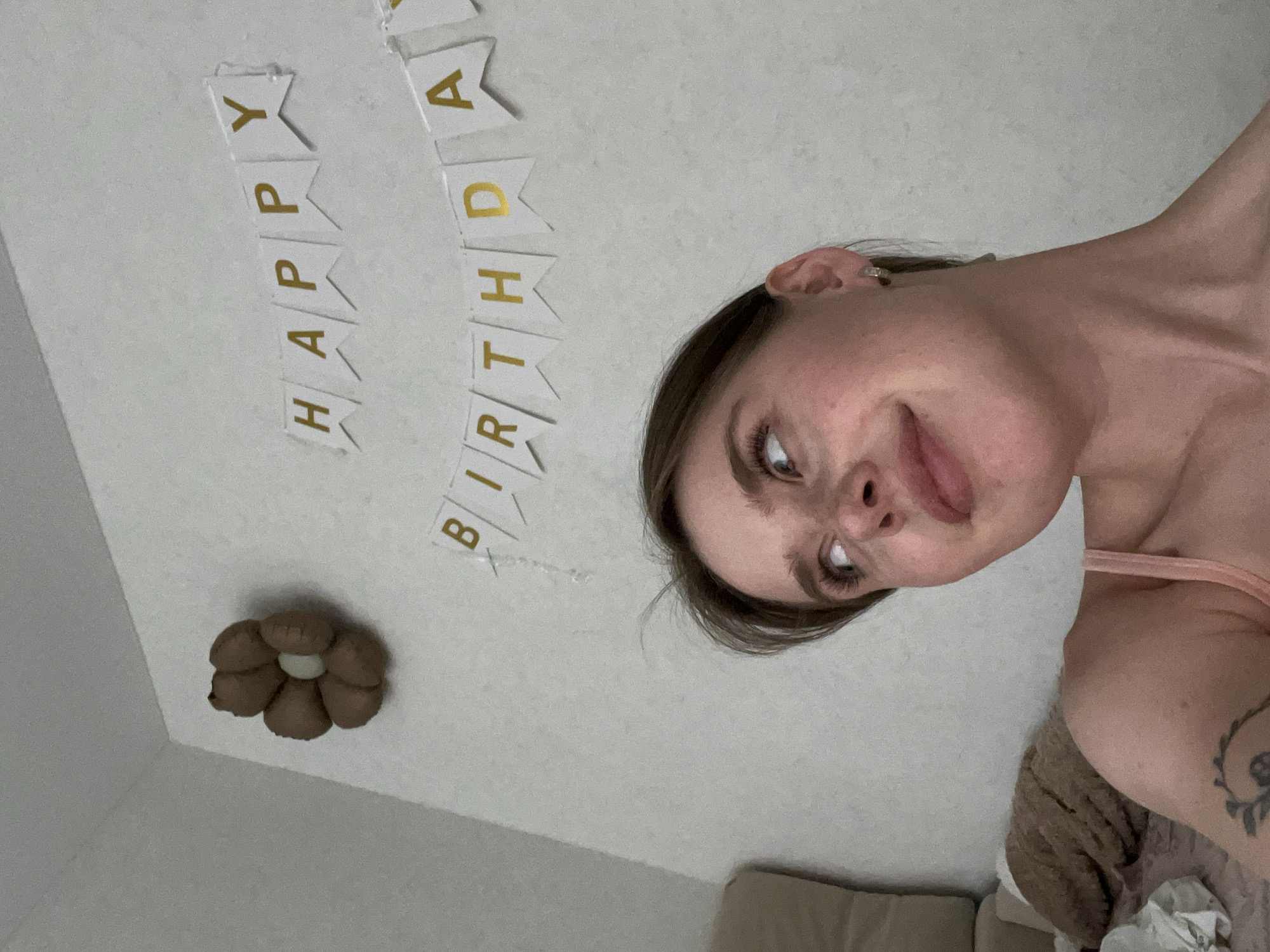 A selfie shows a woman with light skin, brown hair, and visible freckles, positioned in the lower right portion of the frame. She is wearing a pink strap and a partial view of a tattoo is visible on her upper arm. Behind her, a wall is decorated with a "Happy Birthday" banner in gold lettering on white triangular pennants. A brown floral decoration is on the wall to the left of the banner. A beige sofa is partially visible in the lower center and right of the frame. No information suggests it's Chaikovsky, Russia.