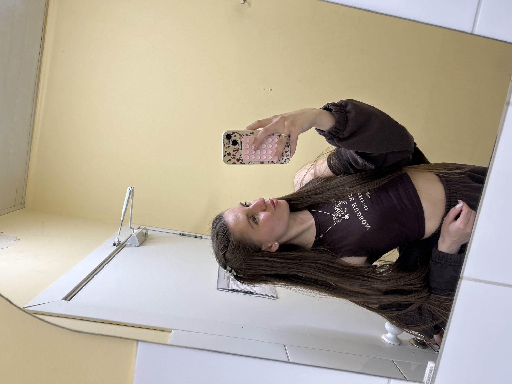 A woman is taking a selfie in a bathroom mirror. She is holding a pink and patterned iPhone. The bathroom has yellow walls and white tiles. A white metal toilet flusher arm is visible to the left of the frame. A toilet paper holder is partially visible in the bottom right of the frame. The woman is wearing a dark brown crop top and a matching zip-up jacket. The text on her shirt reads "HUDSON NICE." Her hair is long and brown, and she is wearing a hair clip on one side. The location is unspecified.