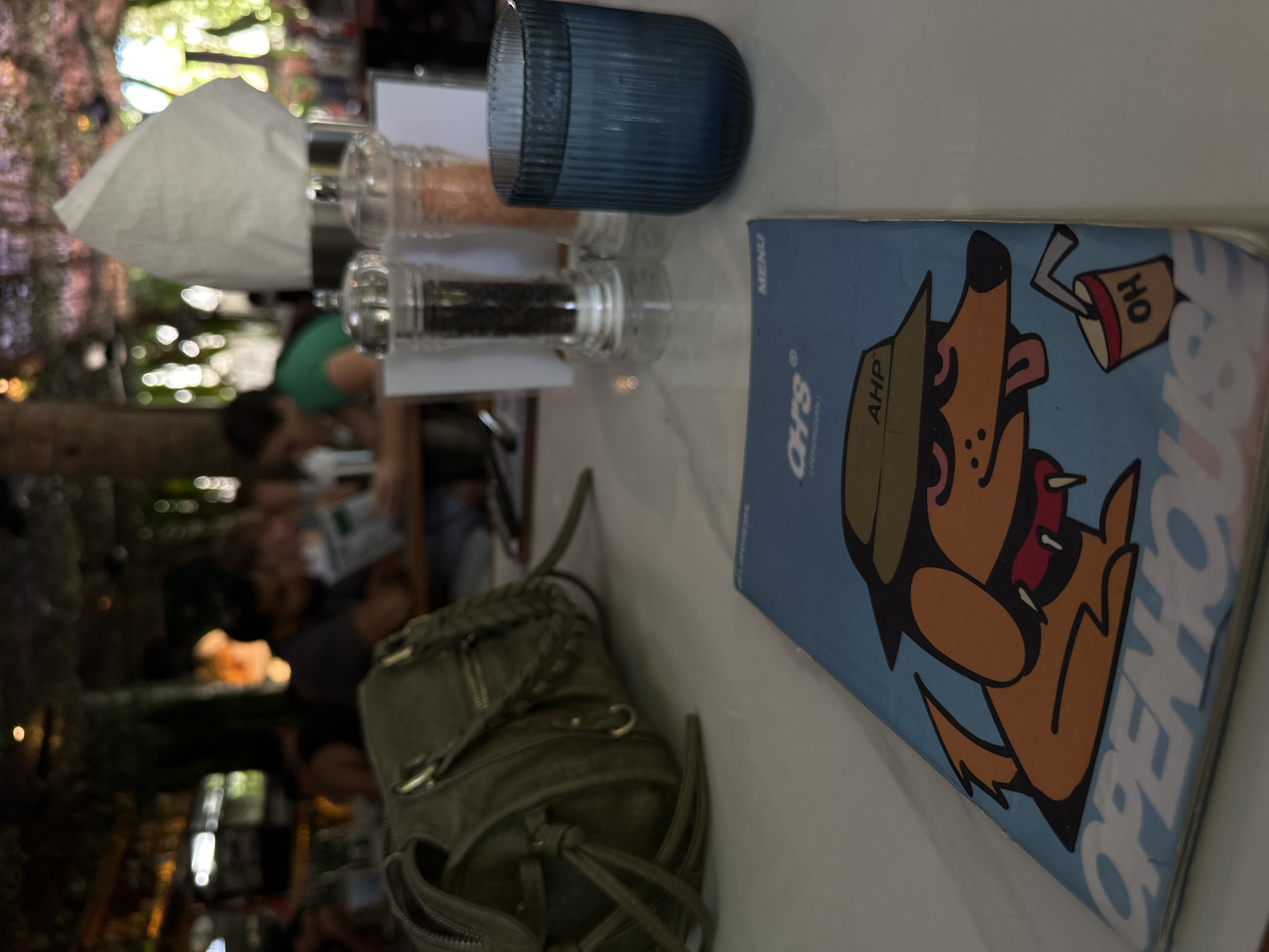 A close-up shot shows a table setting at a restaurant in Mengwi, Indonesia, focusing on a blue menu. The menu features a cartoon illustration of a brown dog wearing a hat and sunglasses, with a red collar and a drink cup that says "OH". The word "OPENHOUSE" is printed in large white letters across the bottom of the menu. In the background, out of focus, are people dining at tables. To the right of the menu, a blue glass filled with water sits next to a salt and pepper shaker set. A green handbag rests on the table to the left of the menu. The environment appears to be an indoor dining area with lush green decorations hanging from the ceiling, suggesting an outdoor-themed or garden setting.