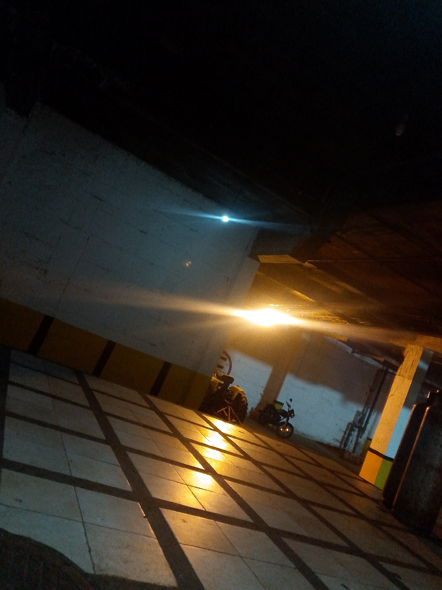 The image depicts an outdoor, paved area, likely a parking lot, at night. The ground is tiled with large, square, gray tiles, creating a patterned effect with dark shadows cast across them from an unseen light source. The walls are concrete, with a strip of yellow and black banding running horizontally along the lower section. There are two motorcycles visible: one larger, yellow and black vehicle is partially obscured by shadow on the left, and a smaller, black motorcycle is parked further back on the right. A bright light source, possibly a streetlamp or vehicle headlights, casts a strong glare across the scene, obscuring details and creating lens flares. The overall impression is one of a dimly lit, utilitarian urban or semi-urban environment. There are no people visible in the image. The context of Pedro González, Venezuela, suggests this may be a public parking area within that locality.