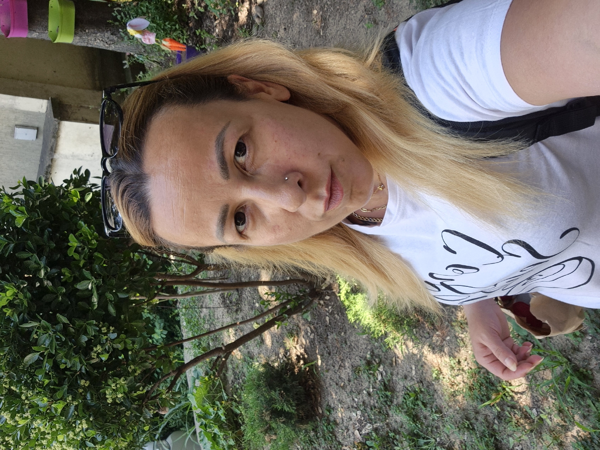 An outdoor, eye-level, medium shot shows a fair-skinned woman with blonde hair, wearing a white t-shirt with a black design, sunglasses perched on her head, and a necklace. She has a nose ring and is holding a brown paper bag. She is standing in a grassy area with trees and bushes in the background. The lighting suggests it is daytime.