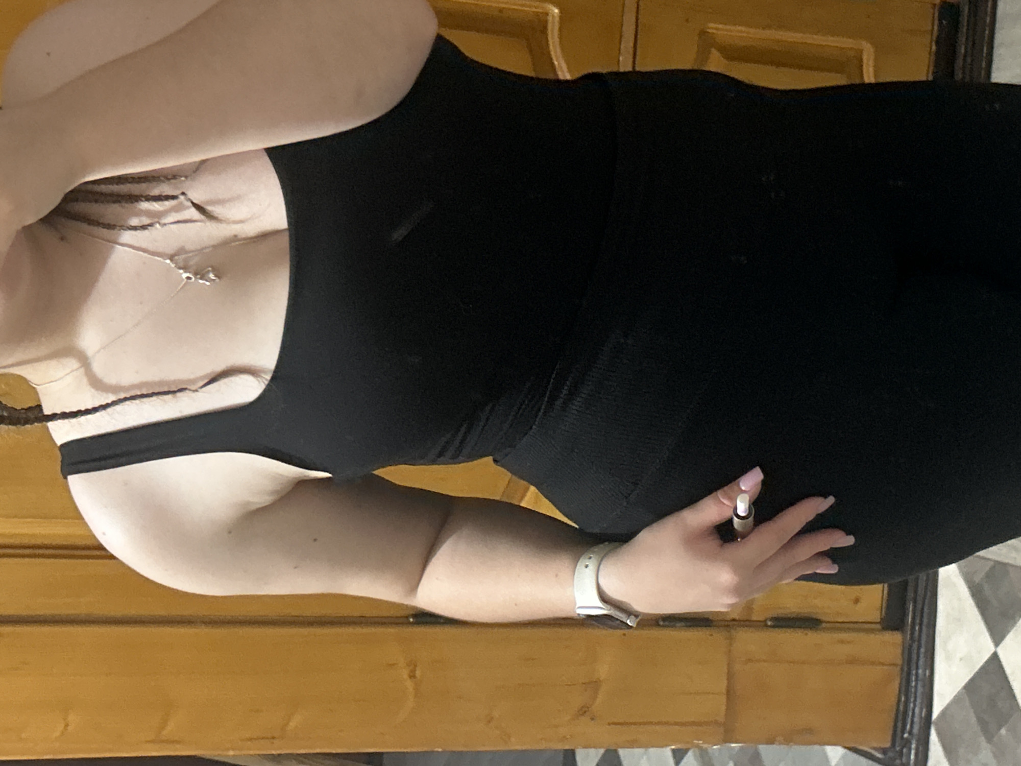 The image shows a close-up view of a person from the chest down, standing in front of a wooden door. The person is wearing a black tank top and black bottoms. A white smartwatch is visible on their left wrist. The person's right hand is holding a small, cylindrical object, possibly a pen or stylus. The background features a light-colored wooden door with vertical panels and a portion of a checkered floor. The scene appears to be indoors. The image does not provide any information about the city of Basarabeasca, Moldova.