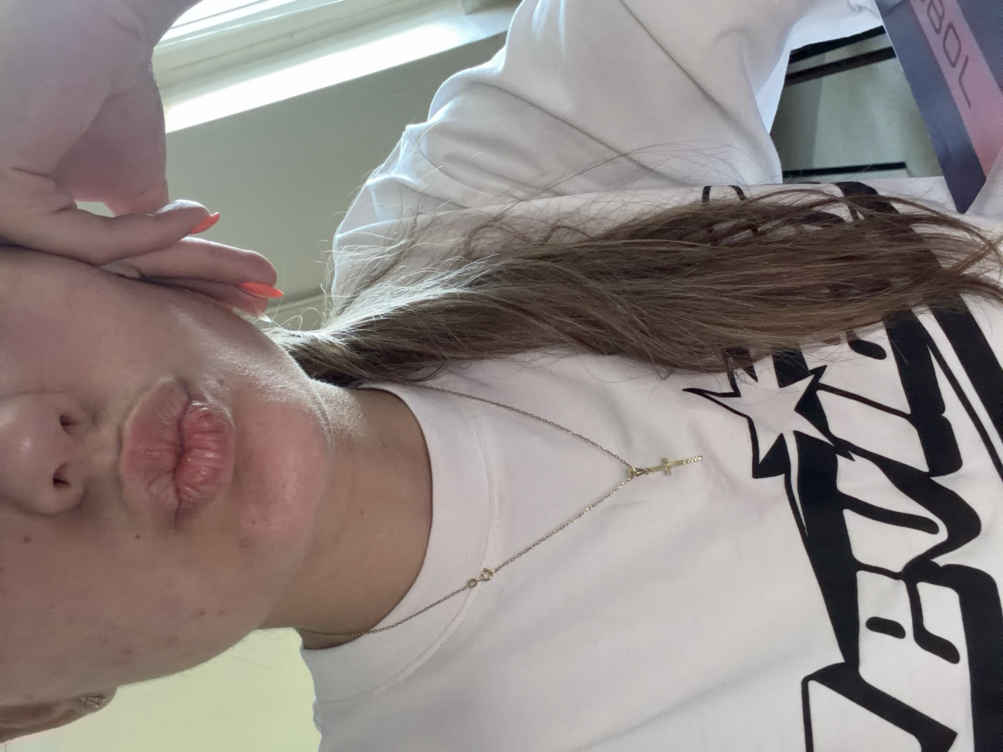 A person with brown hair is captured in an indoor setting, positioned near a window. The person's hand is touching their forehead, and their lips are pursed. They are wearing a white t-shirt with a black design, along with a delicate gold necklace featuring a cross pendant. The background includes a window with blinds, and a pink object is partially visible. There is no information to suggest the image was taken in Burgas, Bulgaria.