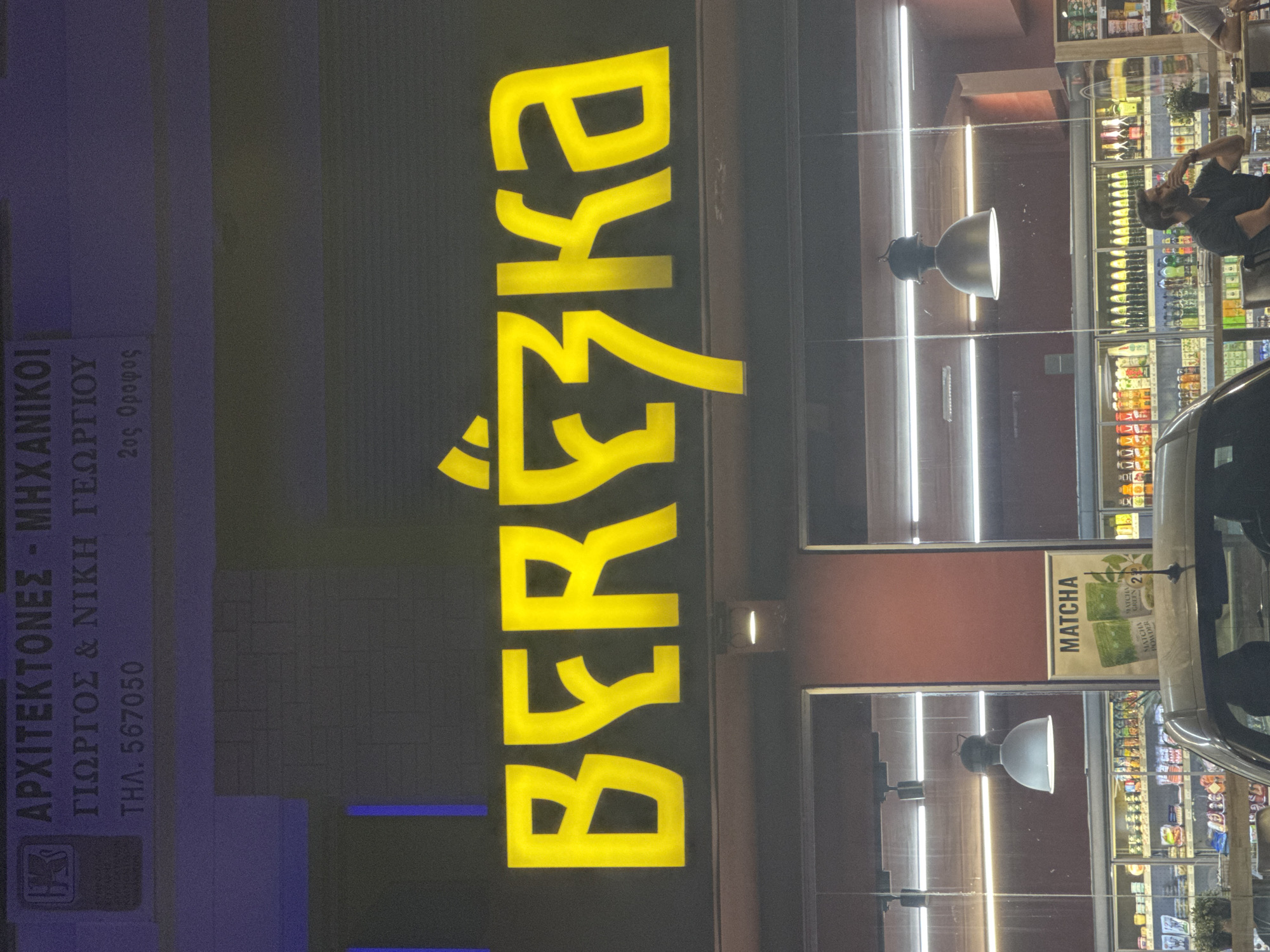 The image shows the exterior of a Bershka store at night, as indicated by the illuminated sign. Above the Bershka sign, there is another sign in Greek that reads "ΑΡΧΙΤΕΚΤΟΝΕΣ - ΜΗΧΑΝΙΚΟΙ ΓΙΩΡΓΟΣ & ΝΙΚΗ ΓΕΩΡΓΙΟΥ ΤΗΛ. 567050 2ος Όροφος". This translates to "ARCHITECTS - ENGINEERS GEORGE & NIKI GEORGIOU TEL. 567050 2nd Floor". The Bershka sign is large, yellow, and illuminated. The store appears to have large glass windows, through which shelves stocked with various products, including beverages and what appears to be a poster for "MATCHA", are visible. A few people are partially visible inside the store. The overall scene suggests a commercial area in an urban setting.