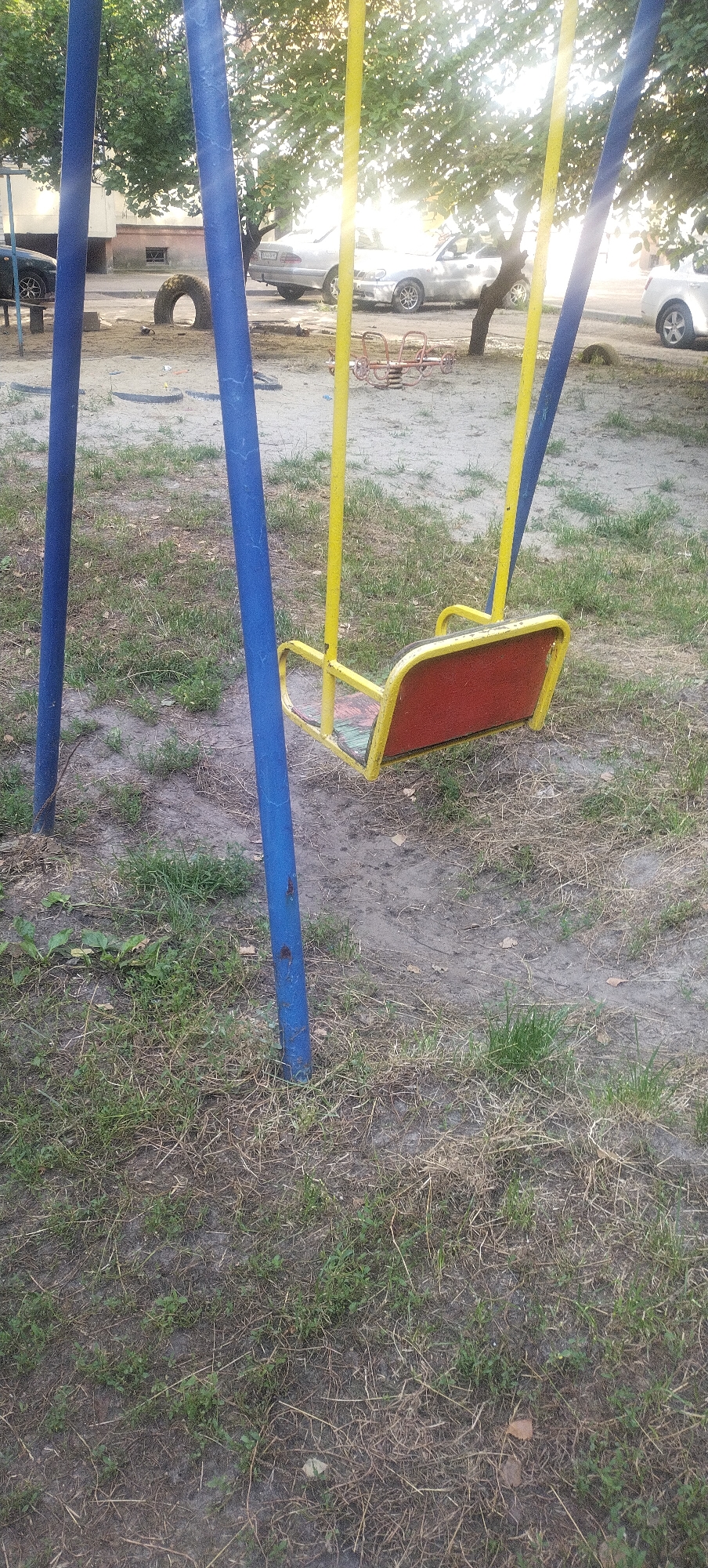 An outdoor scene in the city of Pisochyn, Ukraine, features a yellow and red swing set in the foreground, positioned on a patch of dry grass and dirt. Two vertical blue poles support the swing set. In the background, several cars are parked on a paved surface. A single old tire lies on the ground to the left of the swing set, and a small red metal seesaw is visible further back on the right. Trees with green foliage are scattered throughout the background, and a building with windows is partially visible on the far left. The lighting suggests it is daytime, with sunlight filtering through the trees.