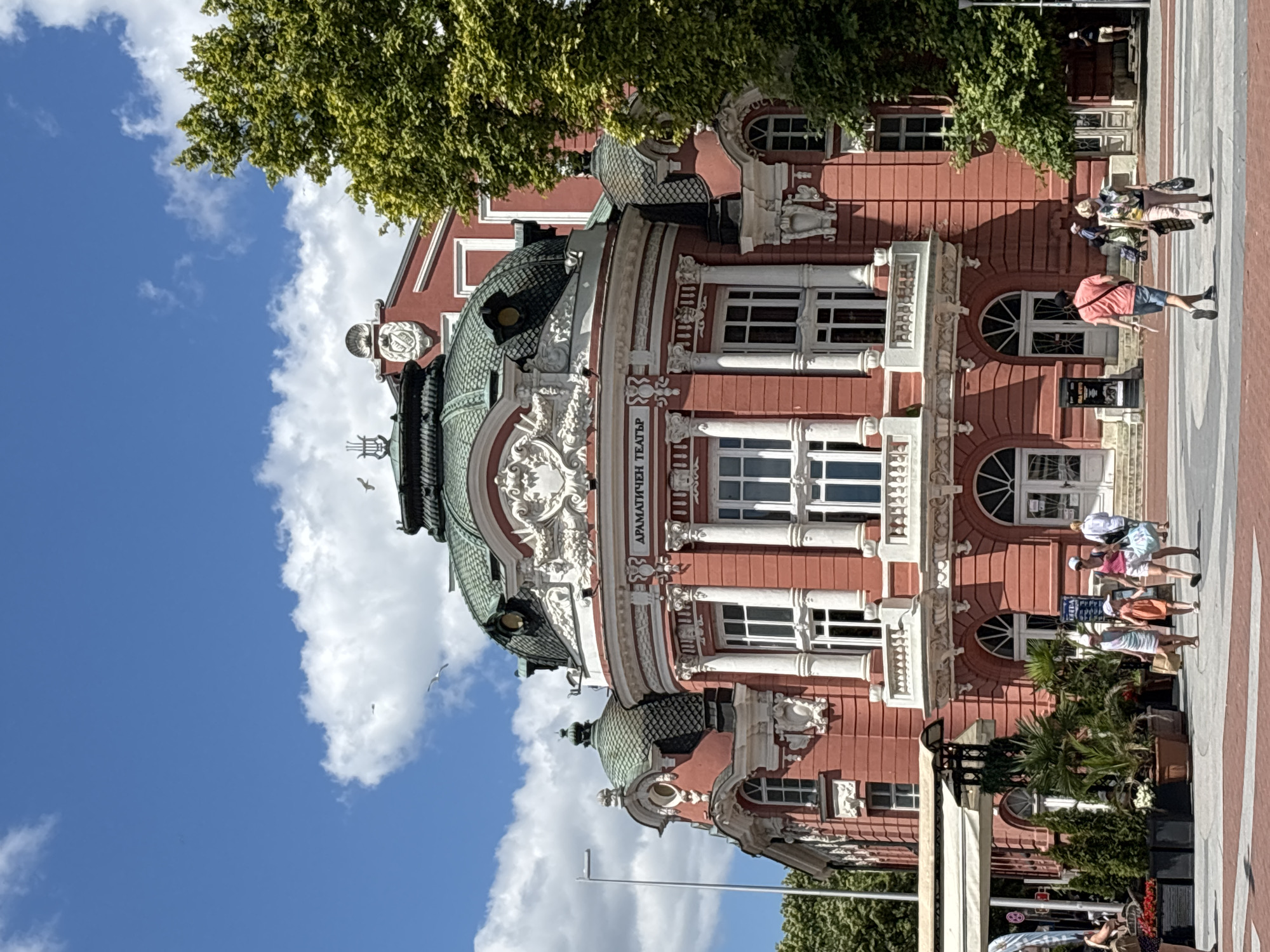 The image captures the exterior of the Dramatic Theatre "Stoyan Bachvarov" in Varna, Bulgaria. The building is predominantly pink with green domed roofs and ornate white architectural details. Several people are walking in the foreground on a paved plaza. One group of three individuals, two women and a child, are walking towards the left. Another woman is walking towards the right, carrying a bag. Further to the right, a man in a pink shirt and blue shorts walks away from the camera. Behind them, a woman in a colorful top and pink pants walks towards the right, carrying shopping bags. The sky is blue with scattered white clouds, and a lush green tree is visible on the right side of the building. Several birds are flying in the sky above the theater. The scene is lit by bright sunlight.